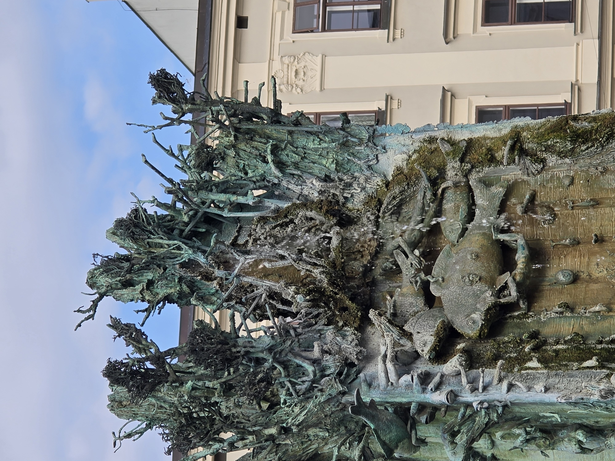 The image shows a close-up of a large, ornate bronze sculpture with a patina of green verdigris, located in the city of Olomouc, Czechia. The sculpture depicts an abstract, organic form resembling intertwined branches or roots, with numerous fish motifs integrated into its structure. Water flows down parts of the sculpture, adding a dynamic element to the scene. In the background, a building with classical architectural features and several windows is visible. The sky above is partly cloudy. This sculpture is likely part of a fountain, as suggested by the water.