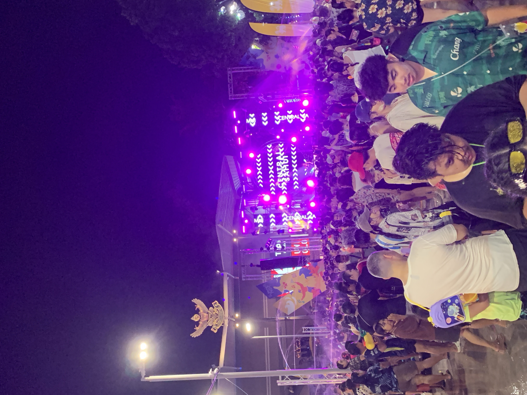The image depicts a large crowd of people gathered at an outdoor event in Baan Chaweng, Thailand. A stage is set up with bright purple lights and banners for "CENTRAL SAMUI". People are seen wearing various clothes and some carrying water guns. The atmosphere appears lively and festive.