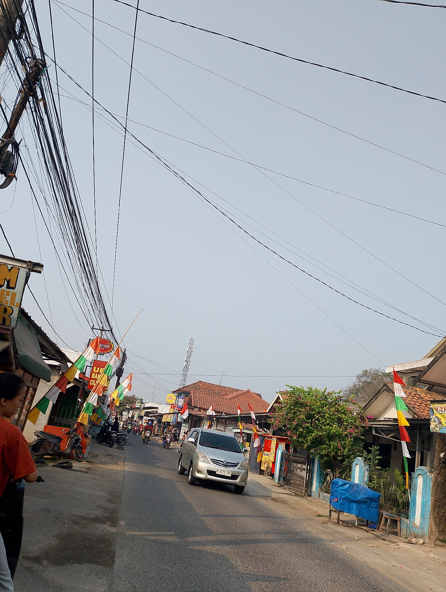The image depicts a street scene in Karawang, Indonesia, likely during daylight hours given the lighting. The street is paved and appears to be moderately busy with vehicular traffic, primarily motorcycles. Numerous overhead electrical wires create a dense network across the sky. Along the sides of the street, there are buildings that appear to be shops or residences, some with awnings. Decorative flags, predominantly in red and white (the colors of the Indonesian flag), are displayed on some of the buildings and poles, suggesting a national celebration or event. A silver sedan is visible in the right lane of the street, while several motorcycles are seen in both lanes and parked on the left side. A person wearing an orange shirt is partially visible on the far left, near a parked motorcycle. The environment shows signs of typical urban or semi-urban Indonesian life, with a mix of infrastructure and local businesses.