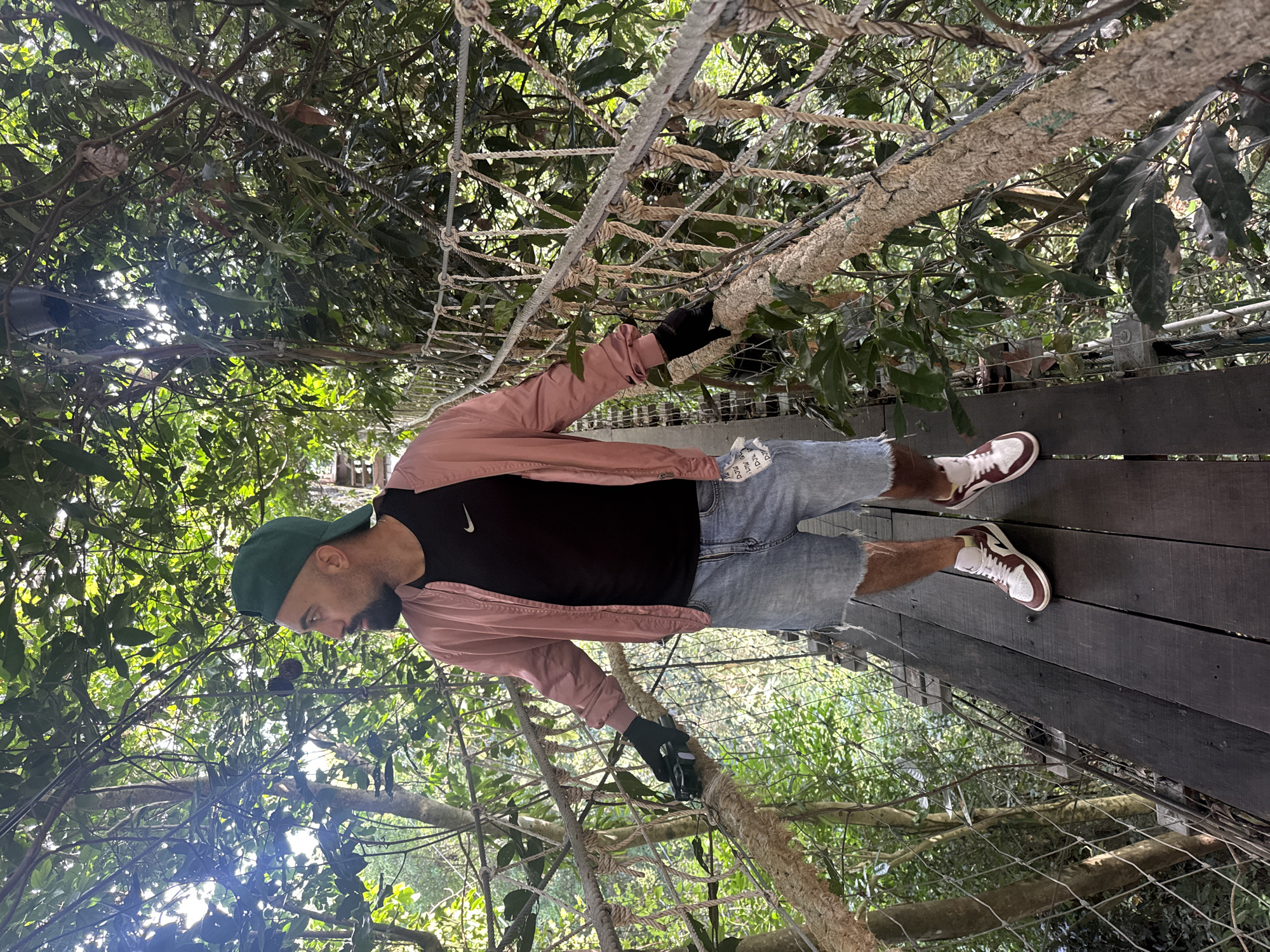 An adult male, wearing a green baseball cap, a pink bomber jacket over a black t-shirt, denim shorts, black gloves, and white and maroon sneakers, stands on a wooden walkway suspended between trees. He is looking to his left and holding onto a rope railing. The walkway is part of a canopy walk, with lush green foliage surrounding it. The scene appears to be an outdoor attraction in Ko Samui District, Thailand, likely a nature park or eco-tourism site. The lighting suggests it is daytime.