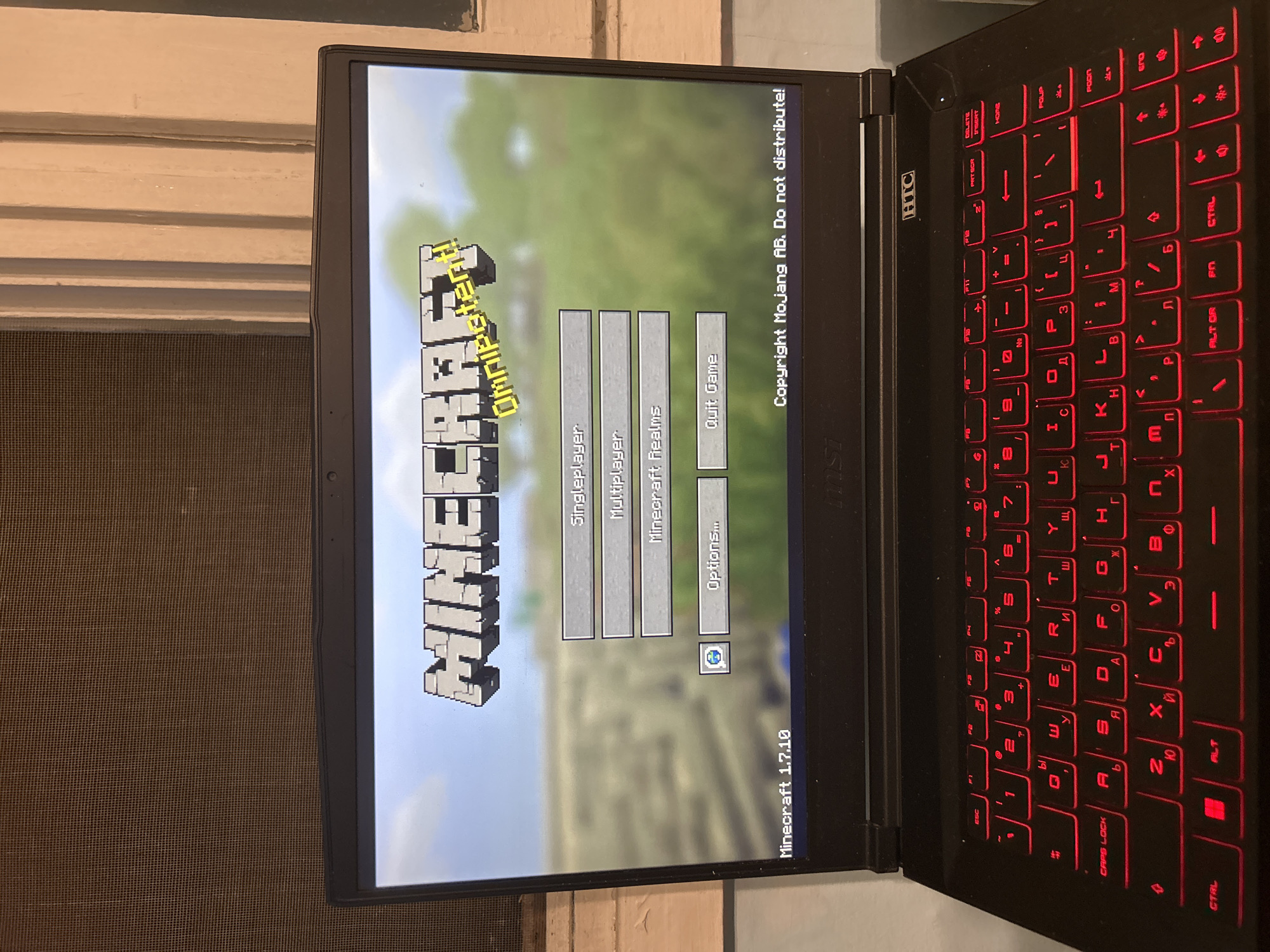 The image shows an open black MSI laptop displaying the main menu of Minecraft version 1.7.10. The options visible are "Singleplayer," "Multiplayer," "Minecraft Realms," "Options...", and "Quit Game." The laptop keyboard is backlit in red, with Cyrillic letters visible alongside the English alphabet. The background shows a window with a screen and white wooden frame.