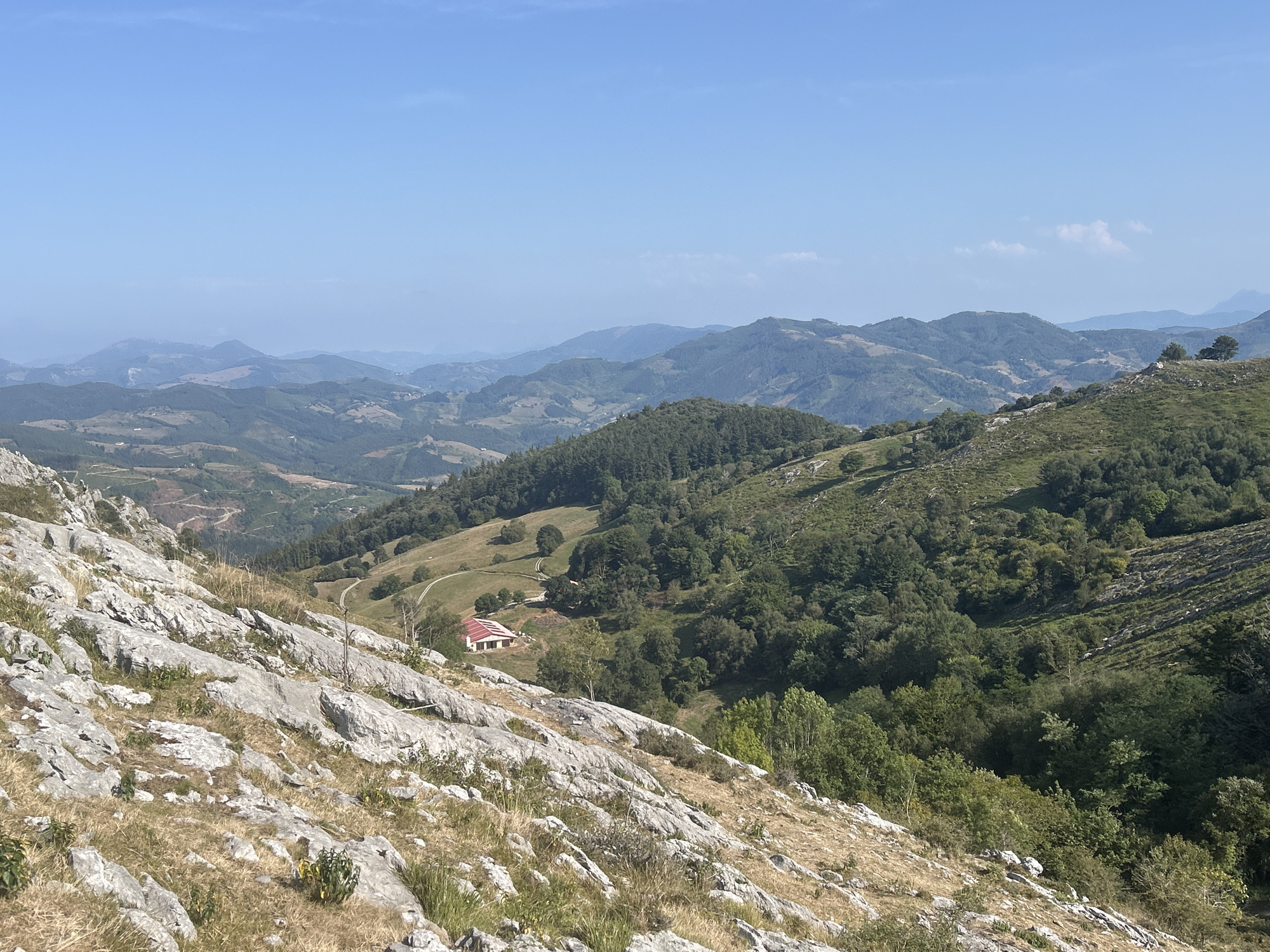 This is a wide, horizontal, eye-level, outdoor, daytime photograph of a mountain landscape. The foreground is dominated by a steep, rocky slope with sparse dry grass and small green plants. Large, pale grey rocks with rough textures are scattered across the slope, suggesting a natural, untamed environment.
In the mid-ground, the terrain descends into a valley. A red-roofed building with white walls, possibly a rural house or farm, is visible on a gently sloping grassy area. A dirt track winds its way towards the building. The valley floor and surrounding hills are covered in lush green vegetation, including dense forests and more open grassy areas. The landscape is characterized by rolling hills and mountains, with layers of peaks receding into the distance.
The background consists of a hazy expanse of mountains under a clear, pale blue sky with a few wispy white clouds. The atmospheric perspective creates a sense of depth, with the farthest mountains appearing lighter and less defined. The image evokes a sense of tranquility and natural beauty. The specific location in Aulesti, Spain, is identified based on contextual information provided with the image, suggesting this is a view from a natural park or hiking trail within the region. No people or animals are present in the frame.
