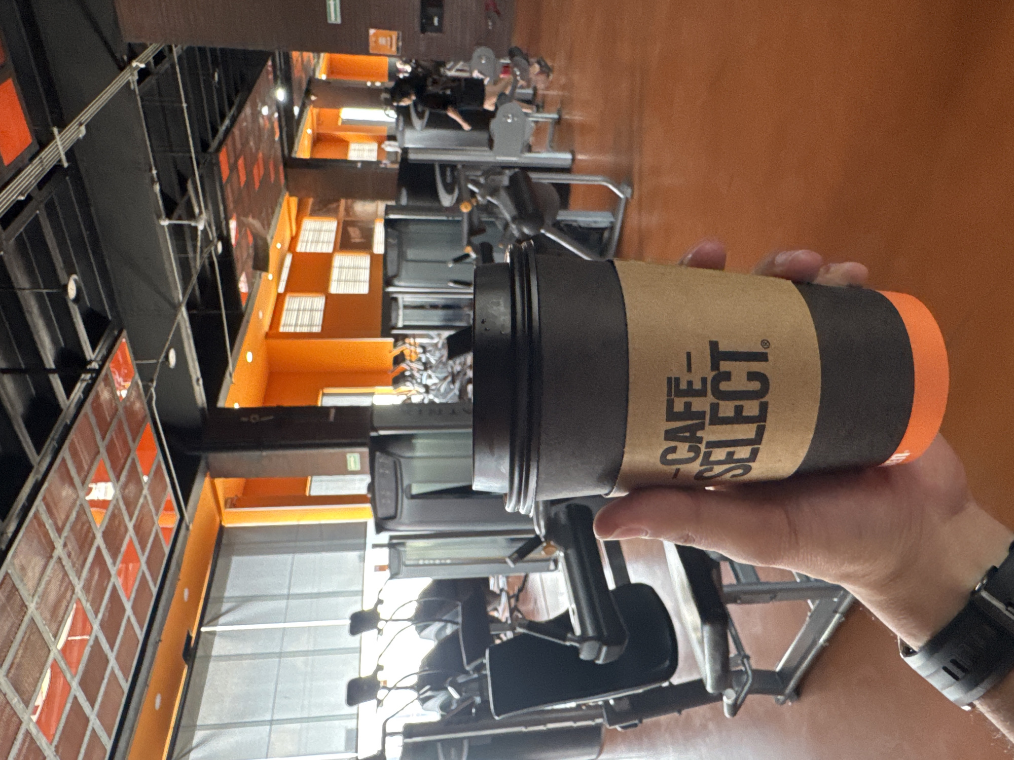 A person holds a "Cafe Select" coffee cup in a gym. The gym is in Puebla, Mexico. There are several people in the background of the gym working out on various weight machines. The gym features orange walls and an open ceiling design with diamond-patterned windows. The person is wearing a black sports watch on their wrist.