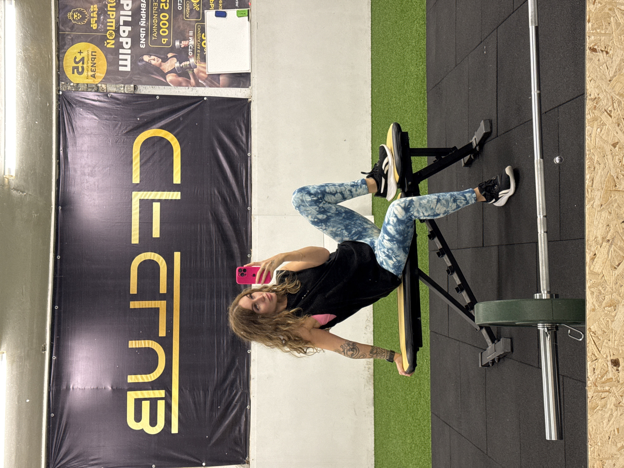 A young woman is taking a selfie inside a gym, captured in a vertically oriented shot that shows her seated on a black fitness bench. She has long, wavy brown hair, wears a black t-shirt, blue and white tie-dye leggings, and black and white sneakers with yellow accents. Tattoos are visible on her left arm. She holds a bright pink-cased smartphone up to take the picture, looking directly at the camera with a neutral expression.
The gym features a modern interior with black rubber flooring, a section of green artificial turf on the left, and a textured light-colored wall. A wooden panel wall is visible on the far right. A silver barbell with a green weight plate rests on the rubber floor.
Behind the woman, two large banners are displayed on the wall. The lower banner is black with prominent gold letters spelling "CLF CLUB". Above it, another banner features Cyrillic text and images related to fitness. Visible words and numbers include "PILUPIM", "UPMON", "АΕΝΟΠ", "+S2", "52.000 b", and "III WECIO". One section of this banner depicts a muscular woman holding dumbbells. The setting clearly indicates a fitness club located in Chaikovsky, Russia, as suggested by the Cyrillic text.