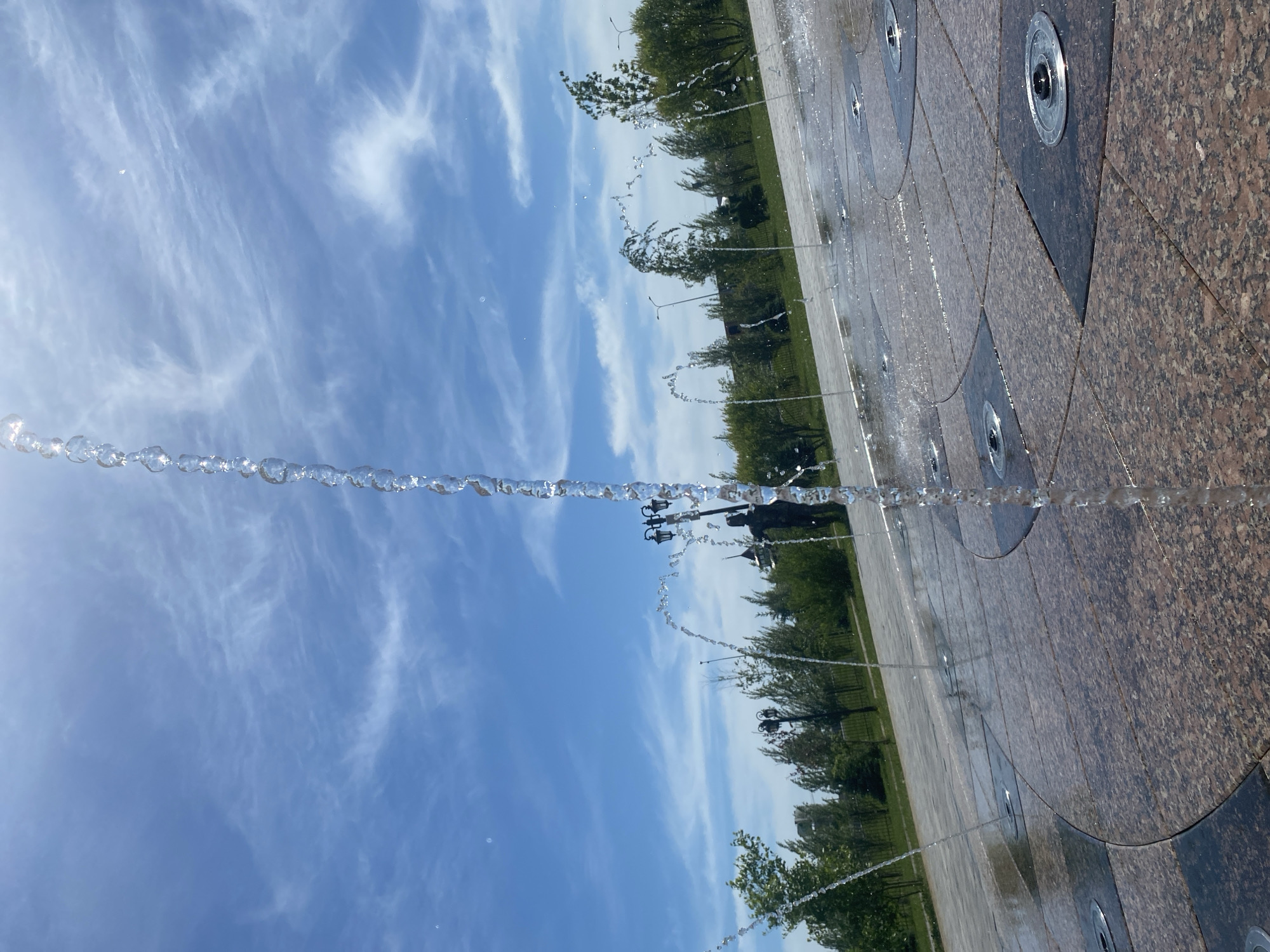 The image displays an active ground-level fountain in a public space in Tyumen, Russia, under a vibrant blue sky dotted with wispy white cirrus clouds. The bright sunlight creates sparkling reflections on the water droplets.
In the foreground and mid-ground, numerous jets of water shoot upwards from nozzles integrated into a paved surface. A prominent stream of water forms an arc across the center of the frame, showcasing distinct, glistening droplets suspended against the sky. The ground is composed of large, polished tiles, likely granite, in alternating shades of brown and dark grey or black, arranged in geometric patterns with circular inlays for the fountain emitters. The tiled surface surrounding the jets is visibly wet.
Beyond the fountain, a lush green area with trees extends towards the horizon. Several streetlights are visible among the foliage, and a dark, indistinct silhouette, possibly a monument or statue, stands in the distance. The scene suggests a clear and sunny day in a well-maintained park or plaza. There are no people or discernible text in the image.