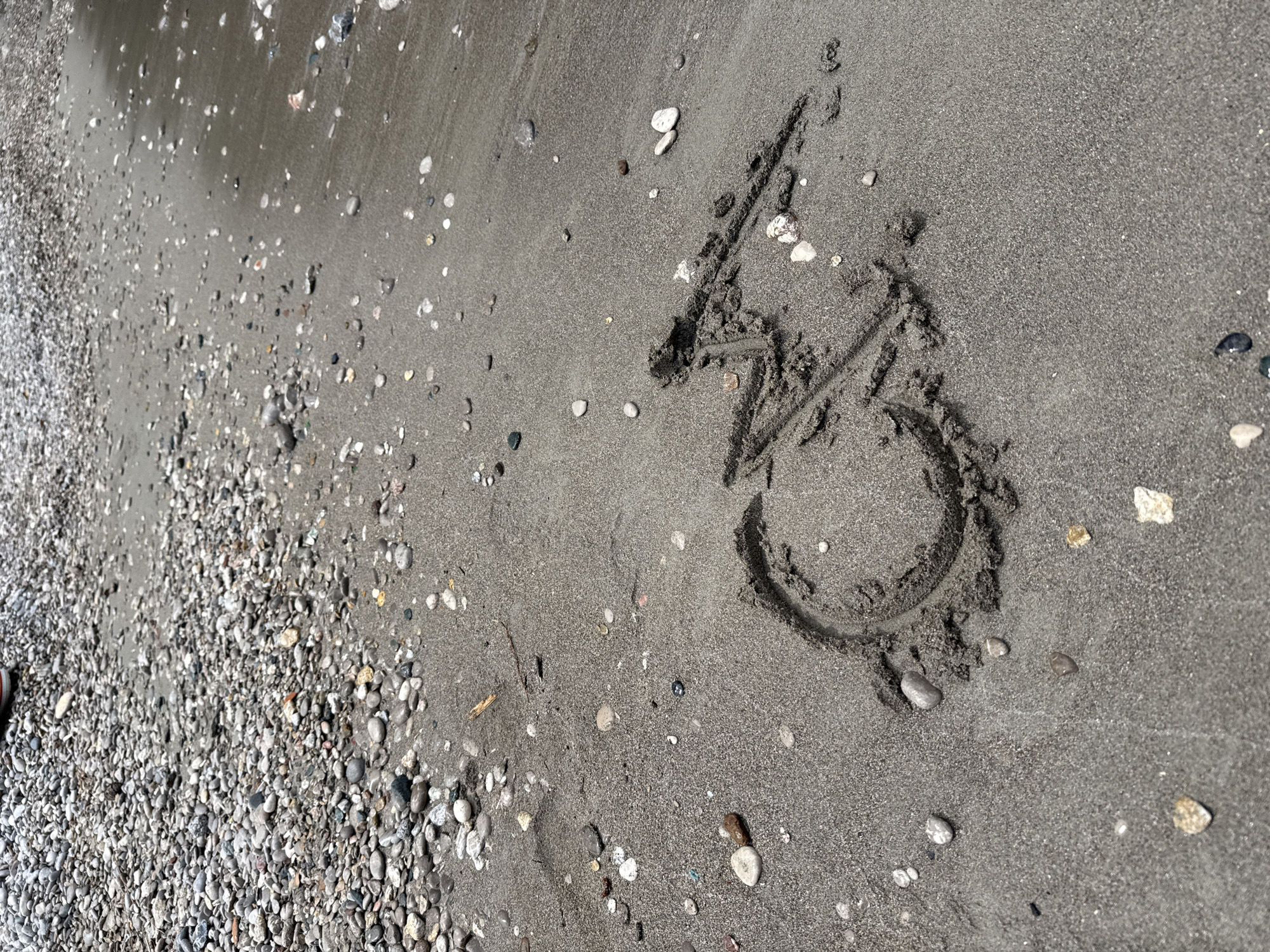 A section of beach in Kumluca, Turkey, shows fine, dark gray sand interspersed with pebbles and small rocks. On the right side of the image, someone has drawn the number "3" in the sand. To the left, there is a higher concentration of small pebbles and rocks that fades into more sand.