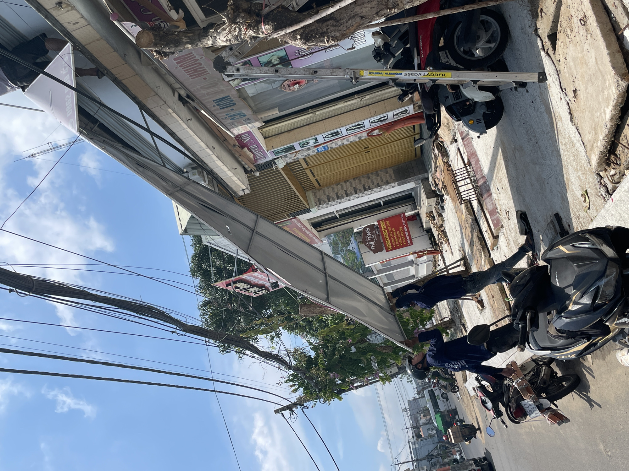 Two men are in the process of installing or removing a large, light-colored panel, possibly an awning or a sign, above a street-level storefront. One man, wearing a dark long-sleeved shirt, dark pants, and a black helmet, is positioned on the left side, looking up at the panel and holding it. The second man, wearing a dark long-sleeved shirt and dark pants, is on the right side, also looking up and assisting with the panel.
The storefront appears to be in a commercial area with multiple businesses. There are signs visible in Vietnamese. One sign on the building reads "CHUYEN: NOI", and another below it displays images of hairstyles. Further down the street, another sign reads "XUONG KHUNG NH...".
The scene is on a street, with power lines and utility poles overhead. Several motorcycles are parked on the side of the street, including a black and gold Honda motorcycle in the foreground and a red motorcycle further back. Construction debris and materials, including concrete slabs, are scattered on the sidewalk and street, suggesting ongoing work or repair. The sky is blue with scattered white clouds, indicating daylight. The overall environment suggests a busy urban or semi-urban setting in Vietnam, possibly in the city of Phu Thuy Ward, Vietnam, given the street signage.