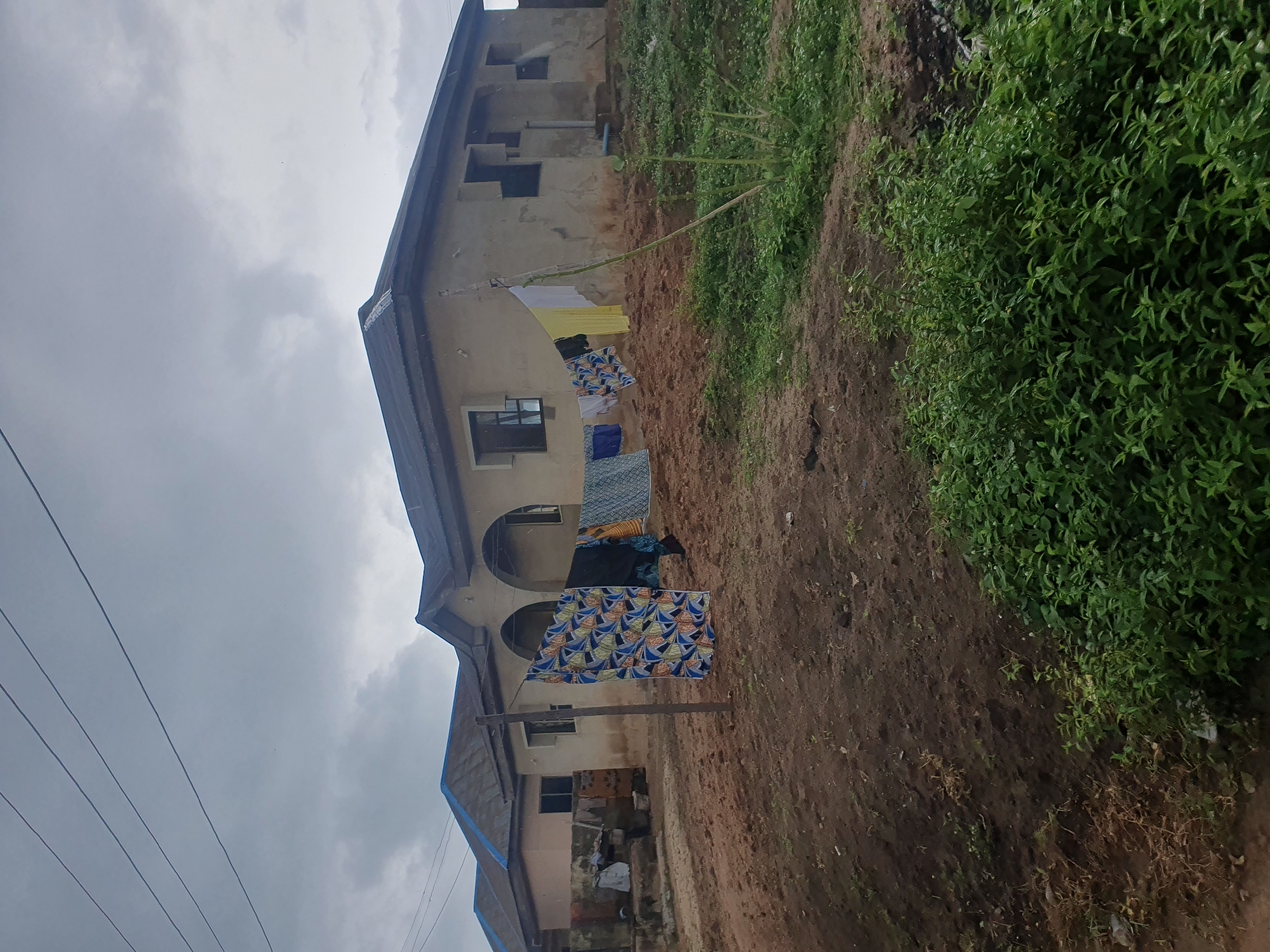 The image depicts an outdoor scene in Lokoja, Nigeria. The primary subjects are buildings and laundry hanging out to dry. The buildings appear to be under construction or in a state of disrepair, with unfinished walls and exposed windows. Several pieces of clothing, including brightly colored patterned fabrics and solid colored garments, are draped on clotheslines strung between wooden poles and attached to the buildings.
The surrounding environment consists of dry, reddish-brown soil with patches of green vegetation, suggesting a rural or semi-urban setting. The sky is overcast with grey clouds, indicating a cloudy or possibly impending rainy weather. The time of day is not explicitly discernible, but the diffuse light suggests it could be morning or afternoon.
Notable details include the presence of electrical wires running across the sky in the foreground, hinting at a populated area. The overall atmosphere is one of everyday life, with the activity of drying laundry being a common sight in many communities. There is no visible text in the image.