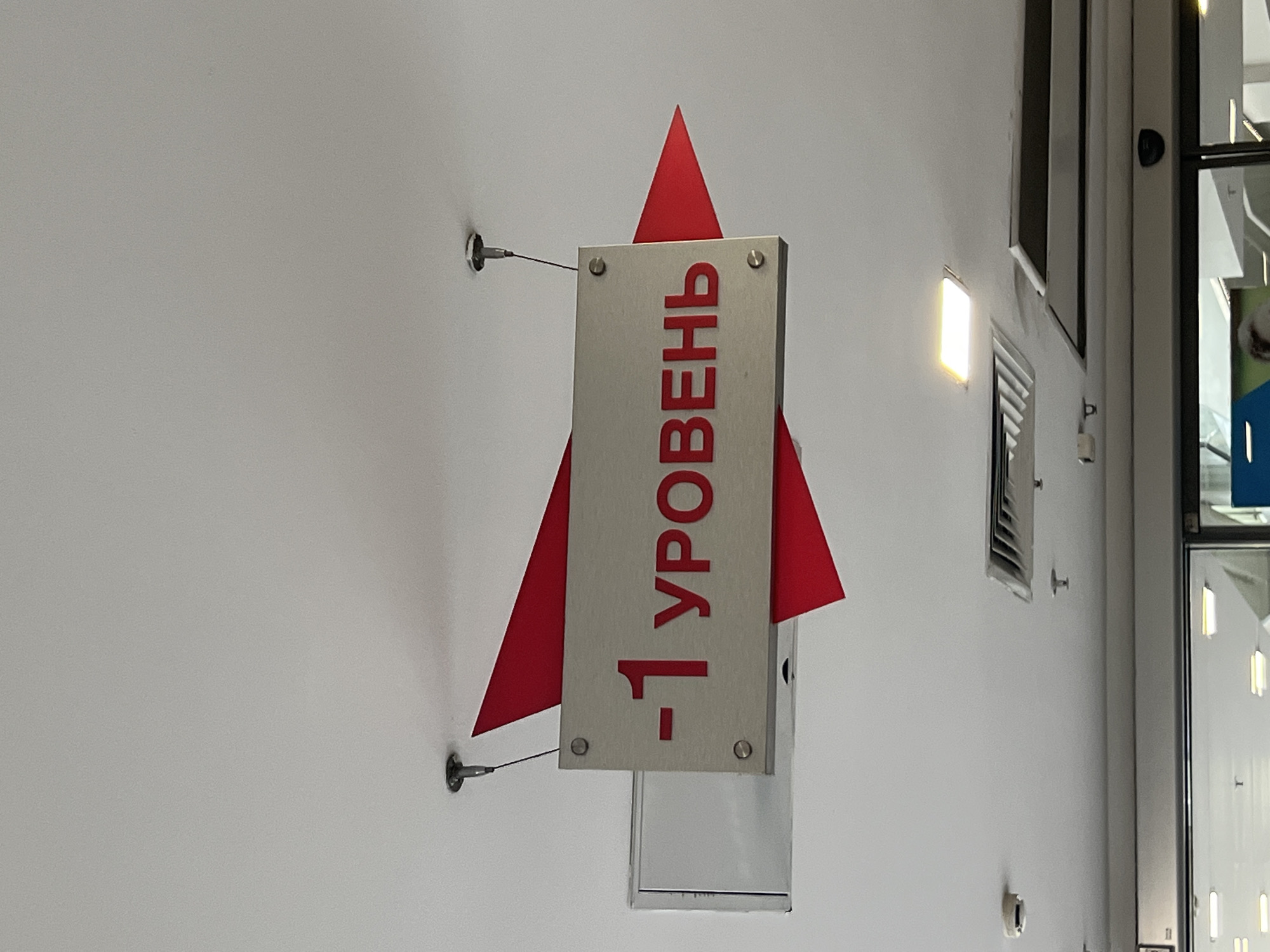 A vertical shot of a sign hanging on a white wall, possibly in a public building in Moscow, Russia. The sign itself is rectangular with a silver-toned surface, attached to the wall by two screws. It is oriented vertically, with text written in red. The text reads "-1 УРОВЕНЬ", which translates to "-1 LEVEL". The sign is designed to resemble a rocket ship, with a red triangular fin extending upwards and two smaller red triangular fins on the sides of the rectangular panel. The rocket design suggests the sign might be indicating a basement level or a lower floor in a shopping center or similar establishment. The image appears to have been taken indoors, indicated by the presence of artificial lighting and interior architectural elements visible on the right side of the frame, including ventilation grates and glass doors or windows. The overall lighting is bright, suggesting daytime, though the specific time of day cannot be determined. There are no people visible in the image.