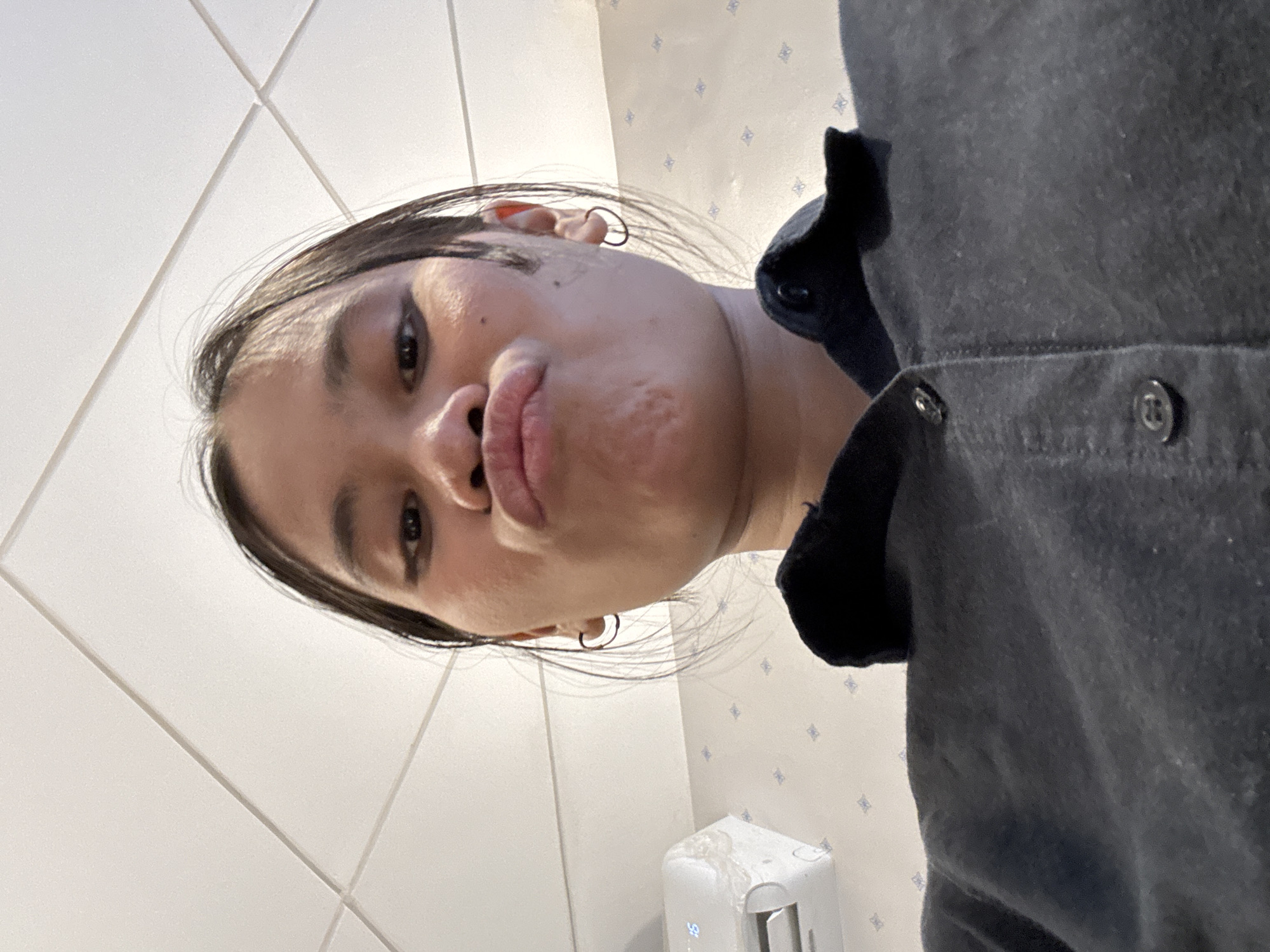 A young woman with dark hair pulled back and hoop earrings puckers her lips for a selfie taken from a low angle. She is wearing a dark button-up shirt with the collar visible. The background consists of a white tiled ceiling and a light-colored wall with a repeating diamond pattern. To the left, a white air conditioning unit is partially visible, displaying the number "16." The overall scene suggests an indoor setting, possibly a room in South Jakarta, Indonesia.