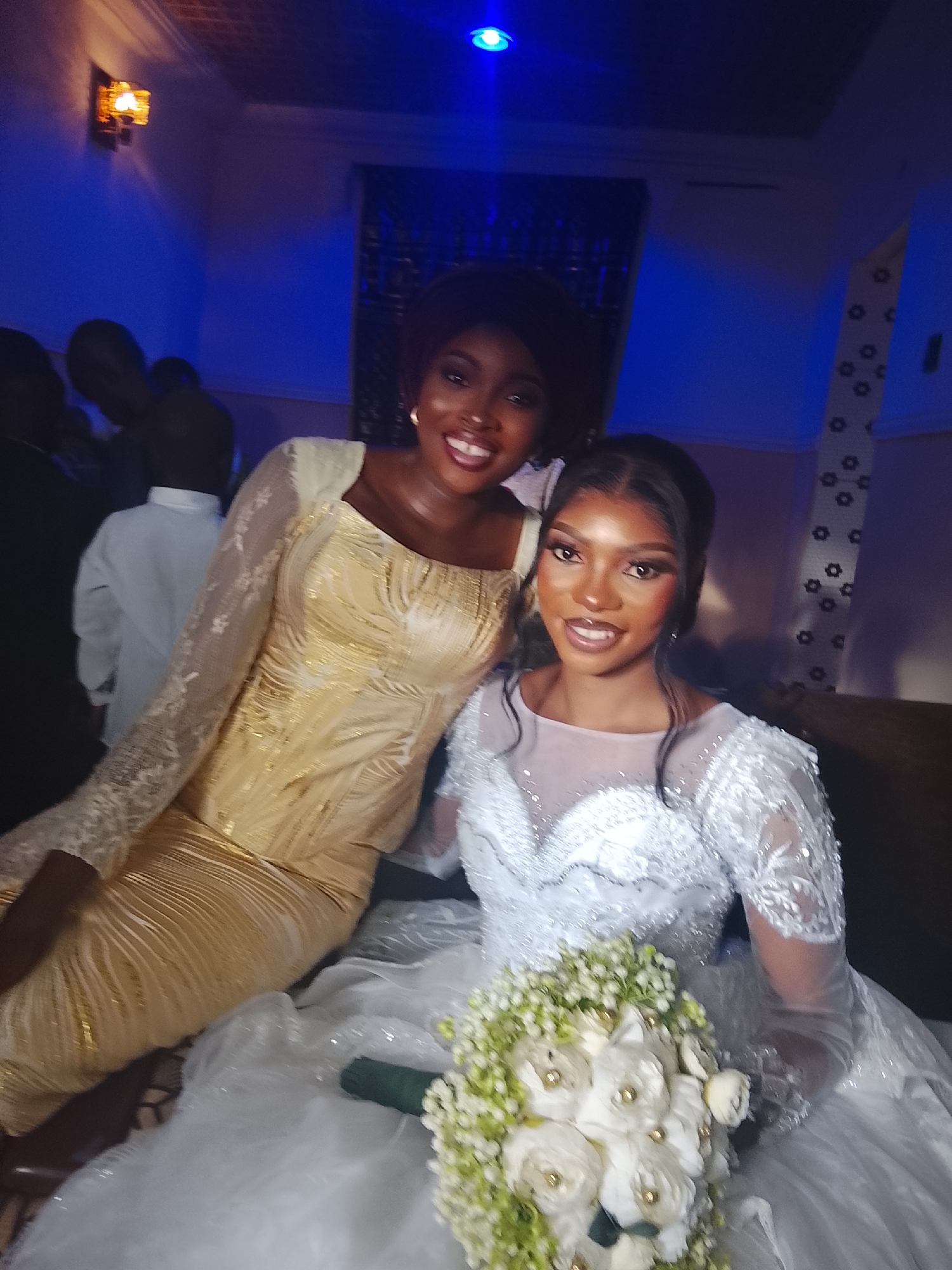 Two women are featured prominently in this indoor photograph, likely taken during an evening event. The woman on the right is presumably a bride, dressed in a voluminous white wedding gown with elaborate lace and beaded detailing on the bodice and long sleeves. She holds a beautiful bouquet of white and light green flowers, some with gold accents. Her hair is styled in an updo with loose strands framing her face, and she wears makeup. She is smiling broadly and looking directly at the camera.
Seated to her left, slightly behind her, is another woman wearing a shimmering gold, long-sleeved dress with a fitted silhouette and lace accents on the sleeves. She also has dark hair and is smiling warmly at the camera. Both women exude happiness and joy.
The background suggests a celebratory setting, possibly a wedding reception or an event hall. The lighting is dim, characterized by deep blue ambient lights from above and a warm-toned wall sconce on the left side of the frame, creating a festive atmosphere. Blurry figures of other guests or attendees can be seen in the background, further indicating a social gathering. No specific text is visible in the image. The overall scene captures a moment of celebration and connection between the two women.