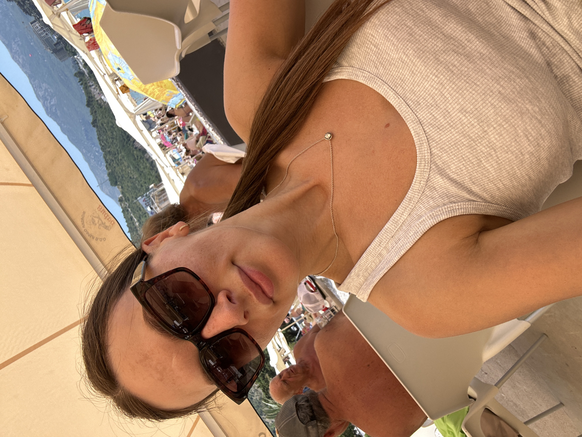 A woman is taking a selfie outdoors, under a beige umbrella, with a beach scene in the background. She is wearing large, dark sunglasses and a white ribbed tank top. A delicate silver necklace with a small pendant is visible around her neck. To her left, an older man with a grey baseball cap is seated, his back to the camera, his bare torso tanned. Further back, other people are visible on a beach with numerous umbrellas and lounge chairs. Mountains form the backdrop against a clear blue sky. The scene appears to be on a beach in Budva, Montenegro.