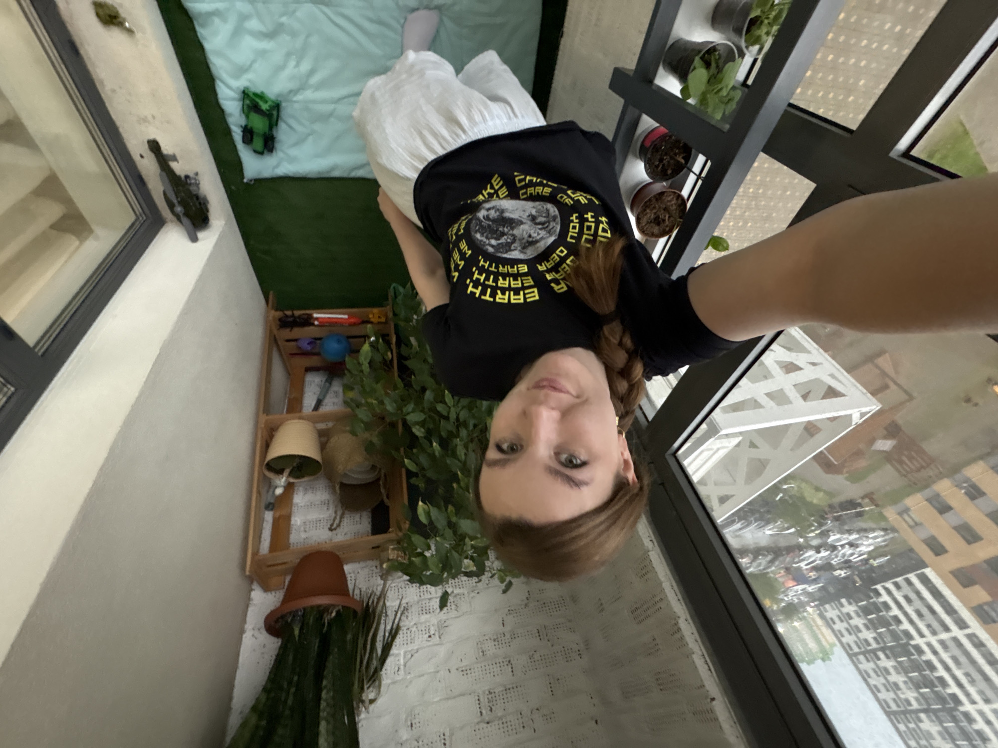 A selfie of a young woman wearing a black t-shirt with an Earth graphic. She is standing on a balcony in Izhevsk, Russia. The balcony has a green carpet, a small mattress, and several plants. Outside the balcony, there are high-rise buildings. There are also small toy vehicles on the balcony.