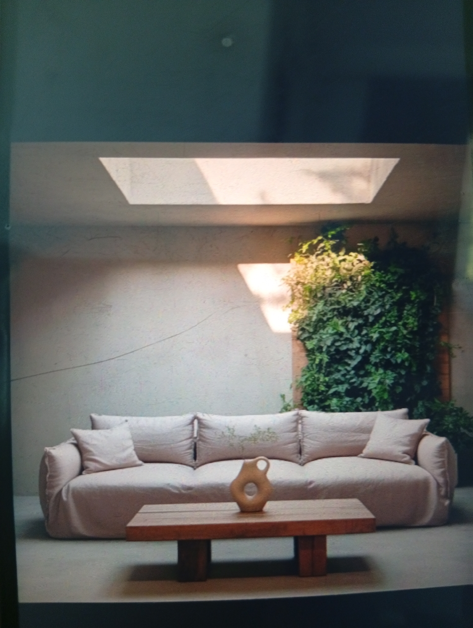 The image shows an interior scene with a large, light pink couch centered in the frame. A low wooden coffee table sits in front of it, holding a ceramic object. Behind the couch is a textured wall with a skylight. To the right, greenery extends up the wall, suggesting an indoor plant or vertical garden. The environment is minimalist. The location of Dutsen Alhaji, Nigeria cannot be confirmed from the image. There are no people visible.