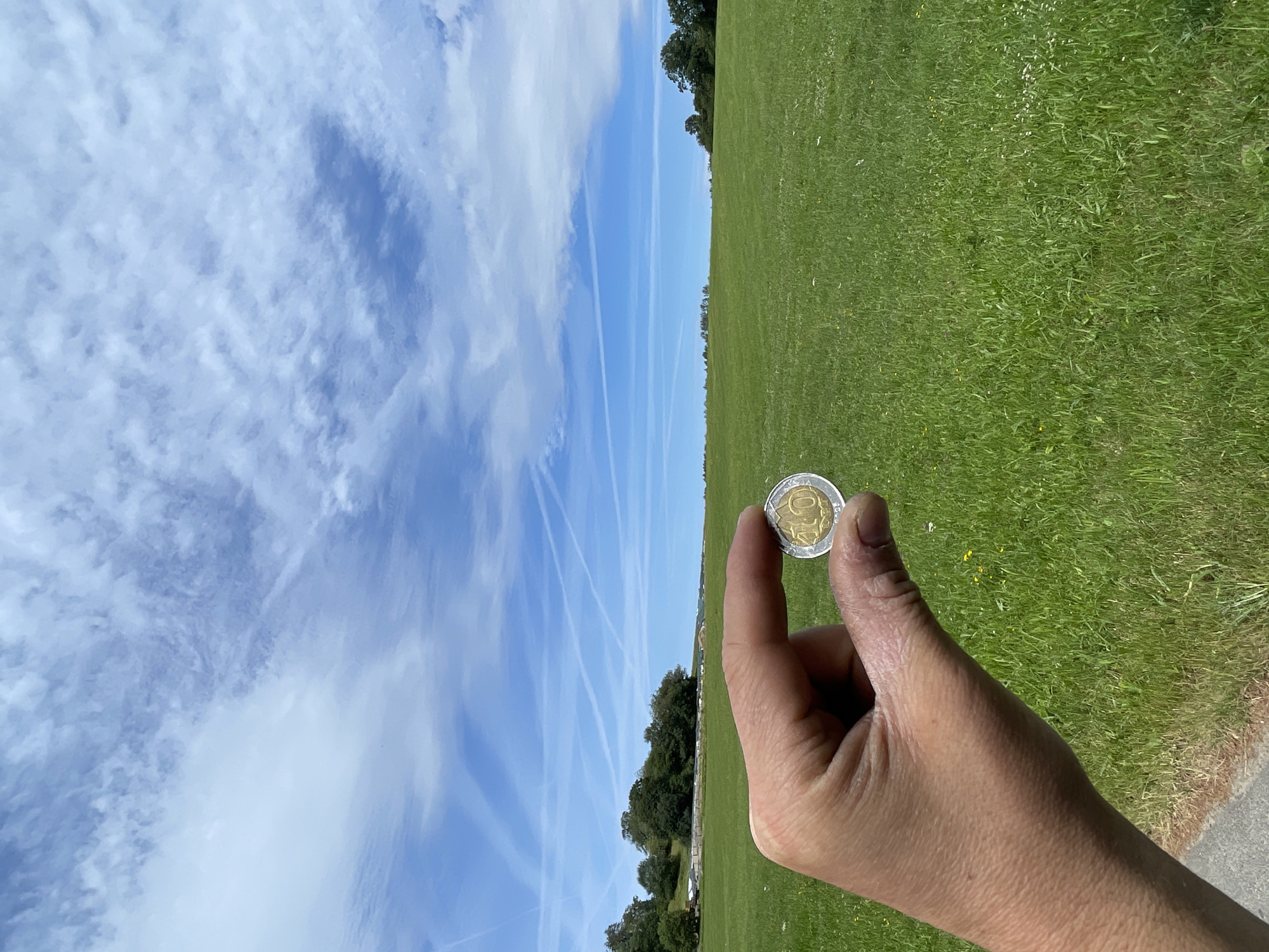 A hand holds a 10 Euro coin in the foreground against a backdrop of a vast green field under a bright blue sky with wispy clouds. The field appears to be a grassy pasture, possibly in a rural or park-like setting, with a line of trees in the distance. The coin is silver and gold-colored, and the number "10" is visible on its surface. The sky is clear with some contrails suggesting air traffic. The overall impression is one of an outdoor scene on a sunny day, likely in the countryside.
