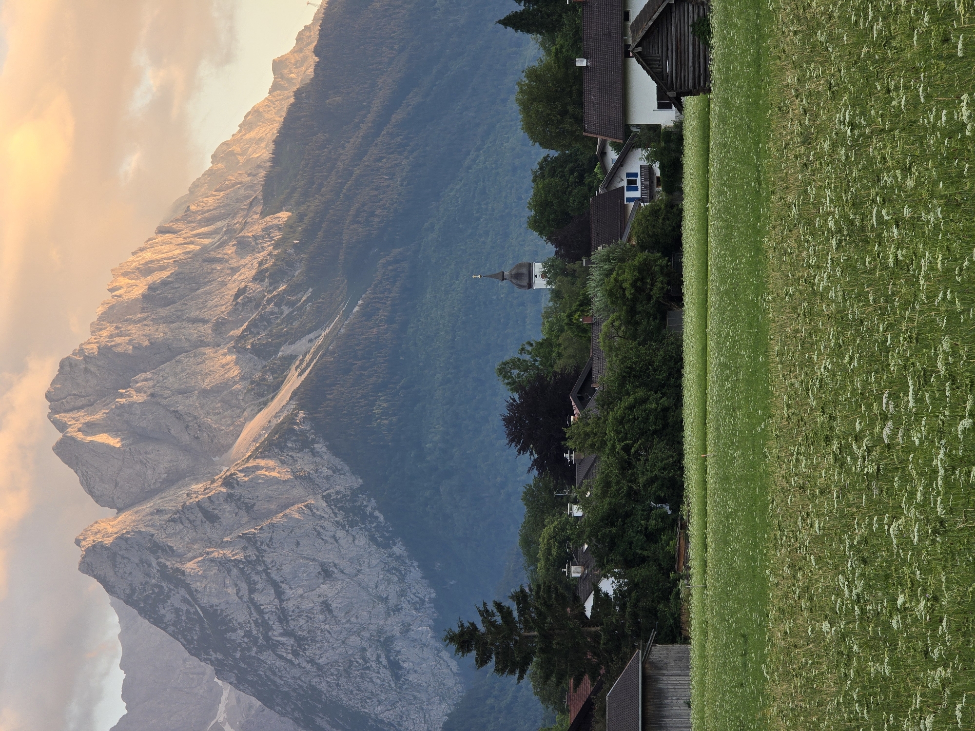 The image depicts a scenic mountain landscape in Garmisch-Partenkirchen, Germany, bathed in the warm light of either sunrise or sunset. The orientation of the image is vertical.
On the left side of the frame, imposing, rugged mountains dominate the scene. Their rocky surfaces are illuminated by the golden hues of the setting or rising sun, casting long shadows and highlighting their textured terrain. Wispy clouds drift across the sky, tinged with the same warm colors.
To the right of the mountains, a dense, dark green forest blankets the lower slopes. Emerging from the treeline is a small village. Traditional Bavarian-style houses with dark roofs are nestled amongst the trees. A prominent church steeple, topped with a cross, rises above the rooftops, suggesting a community setting.
Further to the right, a vibrant green field stretches into the foreground, dotted with small white wildflowers. This field appears to be a meadow or pasture. The overall impression is one of tranquility and natural beauty, with the village harmoniously integrated into the mountainous environment. The time of day is clearly indicated by the dramatic lighting on the mountains and in the sky. There is no visible text in the image.