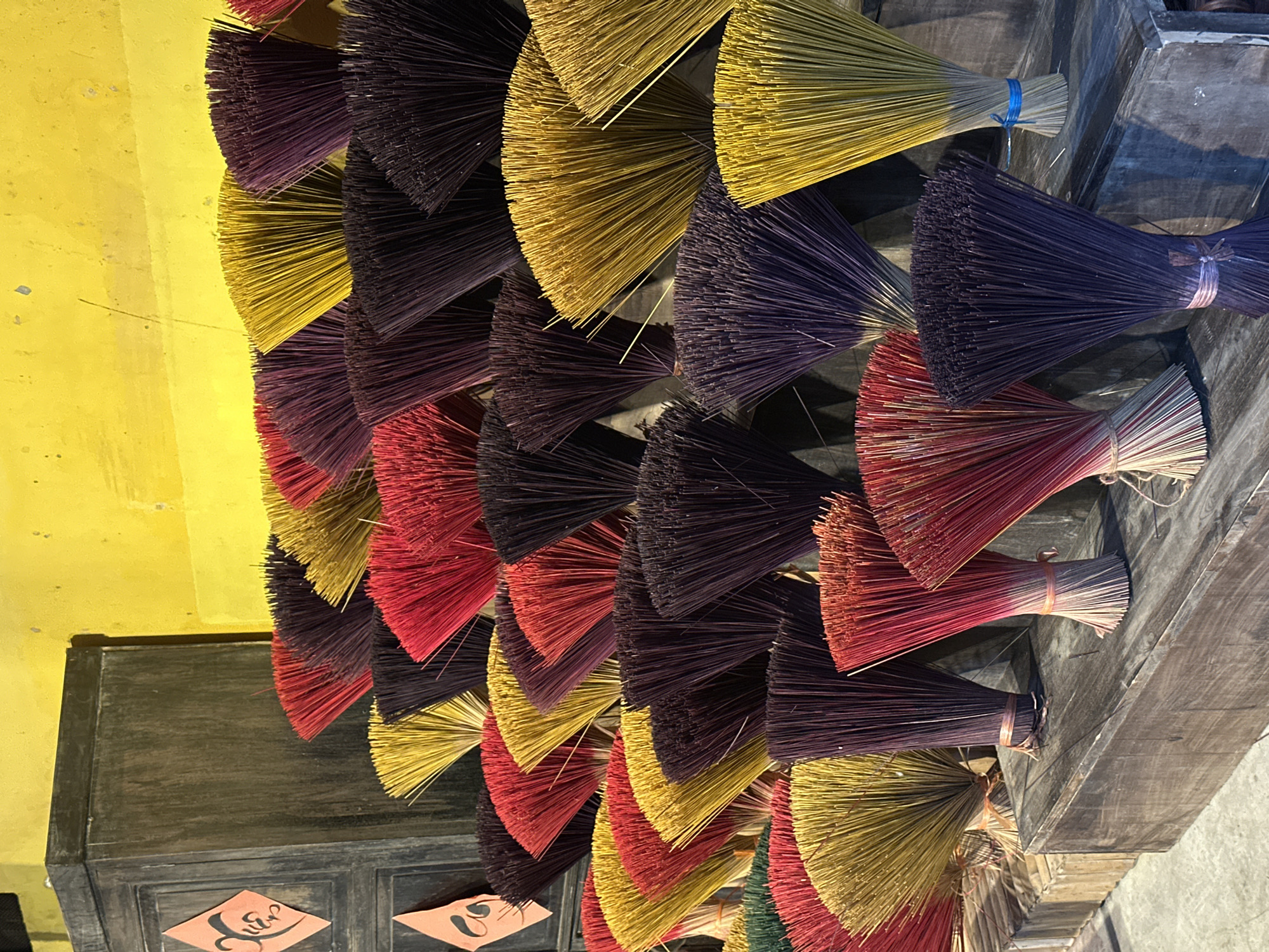 This media file features a close-up, vertical shot of numerous bundles of colorful incense sticks. The subjects are the incense sticks themselves, which are arranged in a fan-like pattern and come in various shades of yellow, purple, and red. They are tied with thin strings or bands. The setting appears to be an indoor space, possibly a workshop or market stall, with a textured yellow wall and dark wooden surfaces visible. Some of the wooden elements show a rustic, aged appearance, possibly forming shelves or a counter.
The activity taking place is the display or drying of these incense sticks. There is no indication of people actively engaged in a task, but the arrangement suggests preparation for sale or use. Notable details include the vibrant colors of the incense, which are neatly organized, creating a visually striking pattern. The lighting suggests it could be daytime, given the brightness of the yellow wall. There are two small orange rectangular tags with some stylized text or symbols on them, attached to the dark wooden surface in the lower left of the frame. One tag has a symbol resembling a handwritten character, and the other has a symbol that looks like a numeral or stylized script. These tags, along with the presence of incense, suggest a cultural or commercial context, potentially related to religious practices or local crafts in Vietnam, given the location cue of Nha Trang. The overall impression is one of vibrant color and organized craftsmanship.