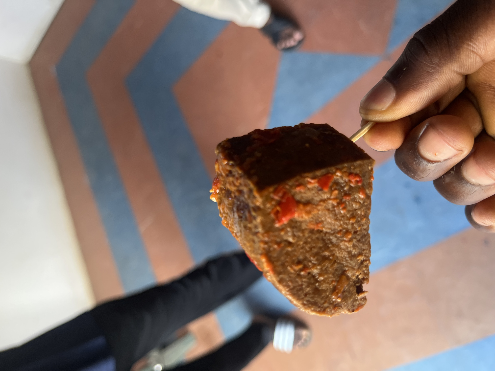 A close-up, slightly rotated image shows a dark-skinned hand holding a brown, cubed meat-like substance impaled on a wooden skewer. The substance appears to have small pieces of red pepper embedded in it. The background features a floor with a chevron pattern alternating between shades of blue and brown. Other elements in the scene include glimpses of two sets of legs and feet; one set is wearing black pants and sandals with white stripes, and the other is wearing white pants and dark-colored sandals. This image was taken in Gura Top, Nigeria.