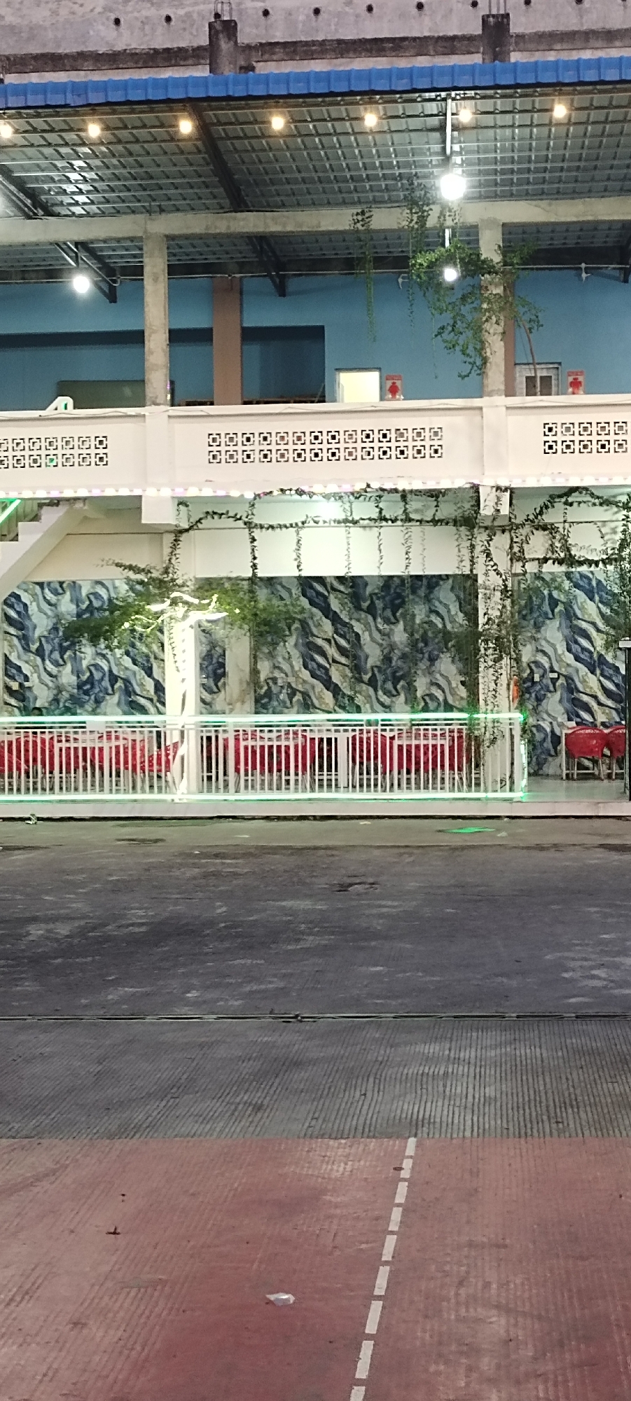 The image shows an outdoor seating area at night in Meunasah Pupu, Indonesia. There are rows of red chairs and tables set up behind a white railing. Vines hang from the ceiling and the wall behind the seating area has a marbled blue and white pattern. The area is lit by overhead lights and green string lights along the railing. The ground in front of the seating area is paved with asphalt and appears to be a walkway or road. There are no people visible in the image.