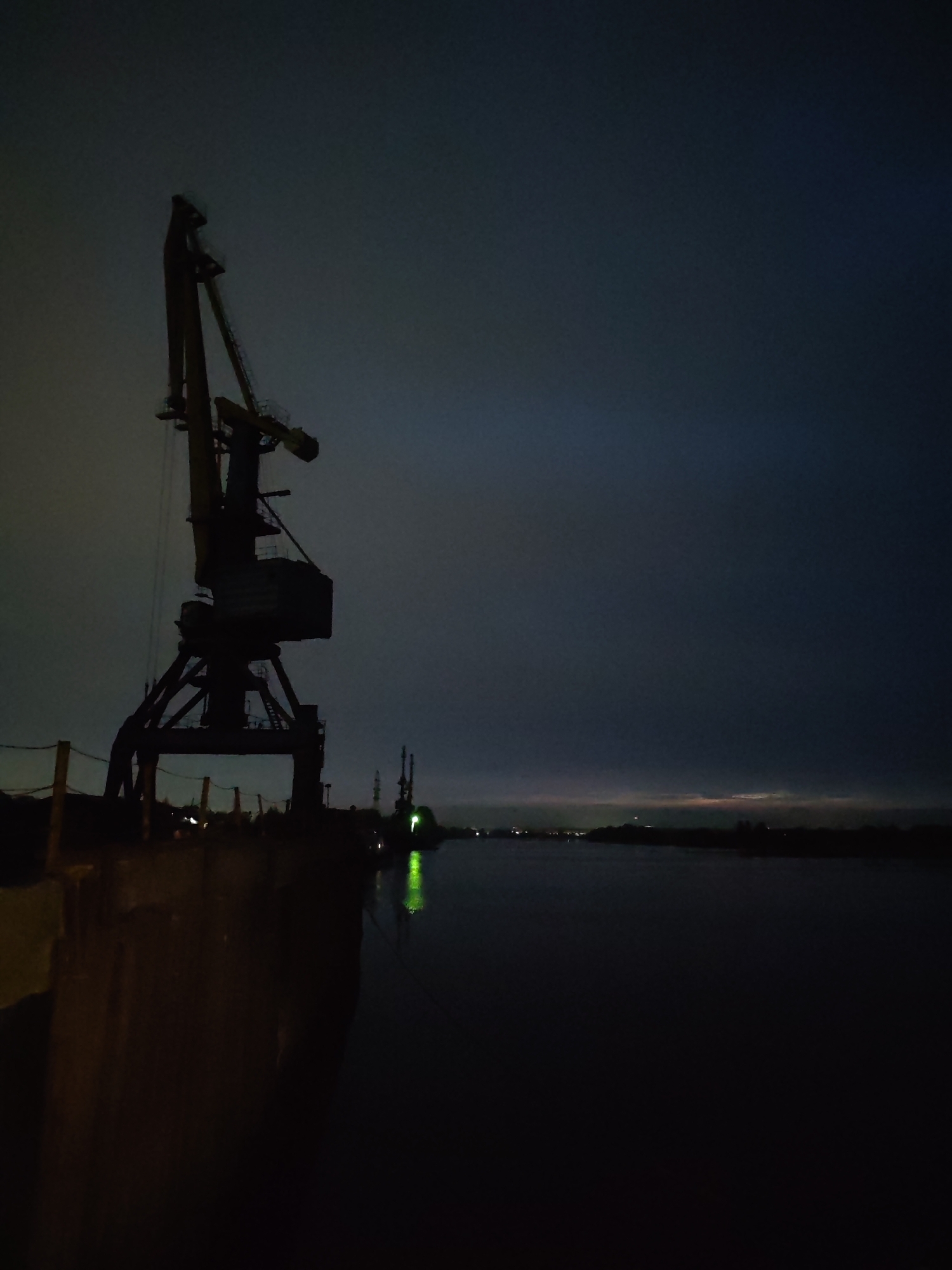The image depicts a dimly lit industrial waterfront scene, likely at night. A large harbor crane is silhouetted against the dark, cloudy sky on the left side of the frame. To the right of the crane is a body of water, reflecting a faint green light from another, more distant object. The cityscape is visible in the background, with faint lights suggesting an urban area across the water. The scene appears to be devoid of people. Given the specification of Tver, Russia, it's plausible that this is a port or industrial area along the Volga River within the city.