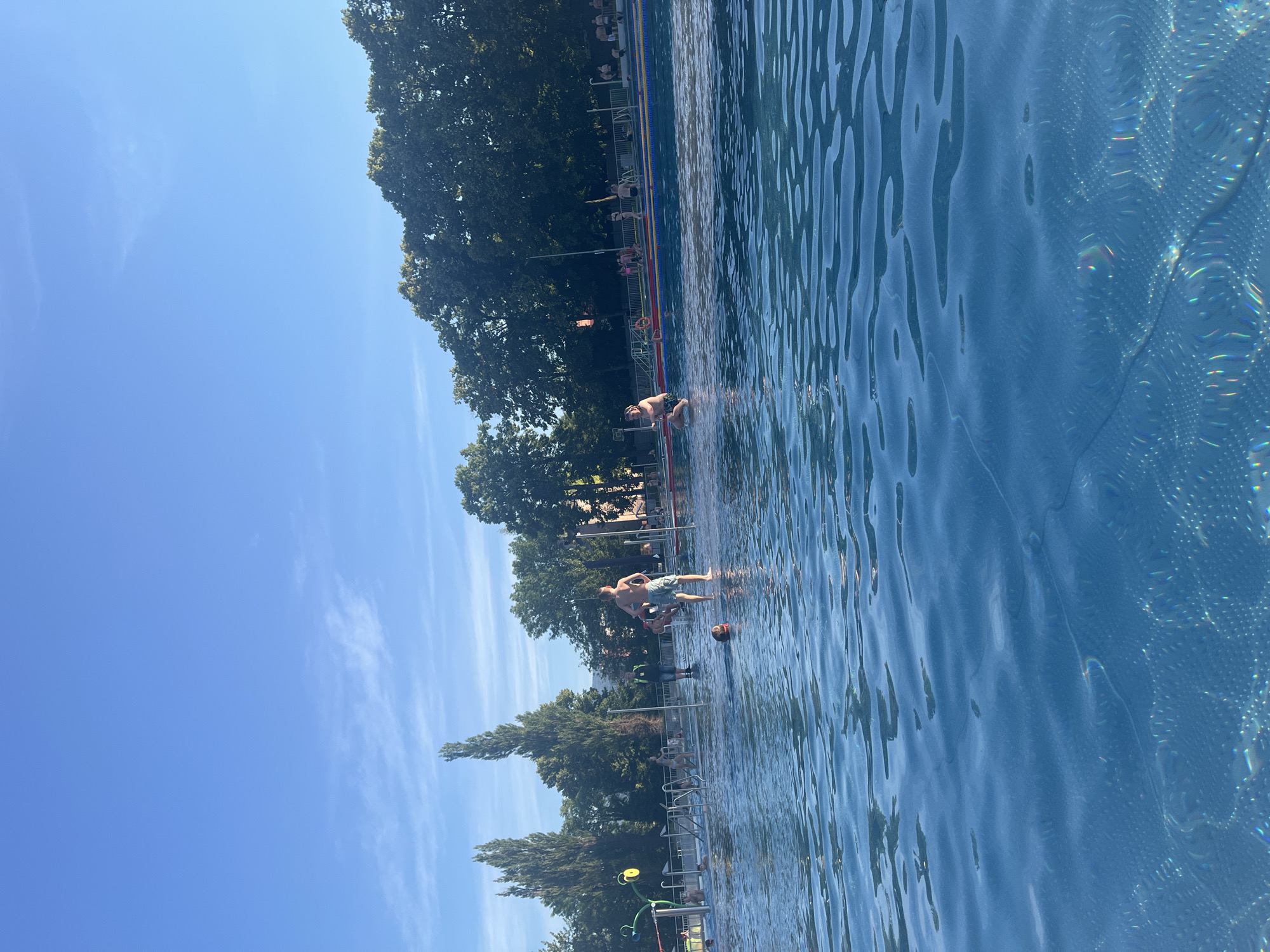 An outdoor swimming pool is depicted under a clear blue sky with scattered clouds. The foreground shows the rippling surface of the water, with light reflecting off it. In the middle ground, several people are visible in and around the pool. Some individuals are swimming or wading in the shallow end, while others are standing on the pool deck. One person, a male, is standing with his hands on his hips, looking towards the water. Another person, a child, is partially submerged in the water. In the background, there are tall trees and buildings. The overall scene suggests a public swimming pool on a sunny day, with people enjoying recreational activities.