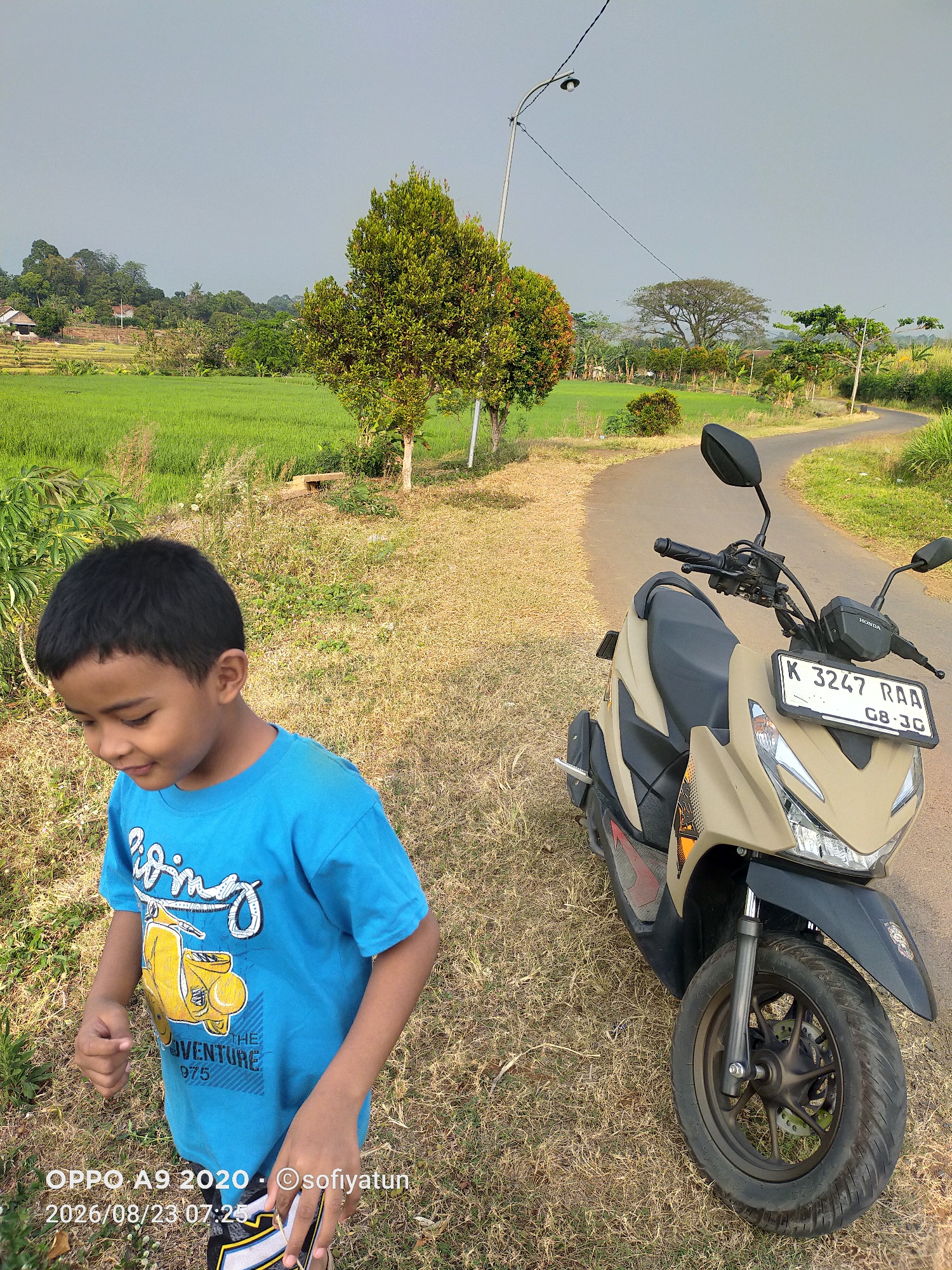 The image depicts a boy in a blue t-shirt standing on a dirt path next to a beige and black scooter. The scooter is parked on the right side of the frame, with its front wheel prominently featured. The boy is in the foreground on the left, looking down and to his left. Behind them, a winding paved road curves through a rural landscape with lush green rice paddies. In the background, trees and scattered houses are visible on rolling hills under a hazy sky. The image is marked with a timestamp indicating it was taken in Tayu, Indonesia, on August 23, 2026, at 07:25.