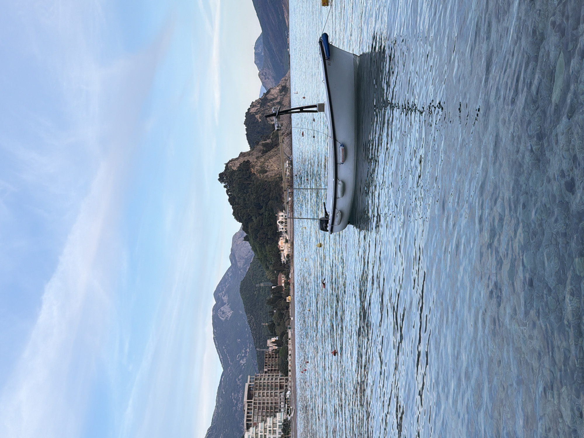 Captured at Petrovac centar, Montenegro, the image showcases a serene coastal scene. A white boat is anchored in clear, rippling water near a shoreline. The backdrop features layered mountain ranges under a sky transitioning from pale blue to a softer hue at the top. A hotel and other buildings are visible near the beach.