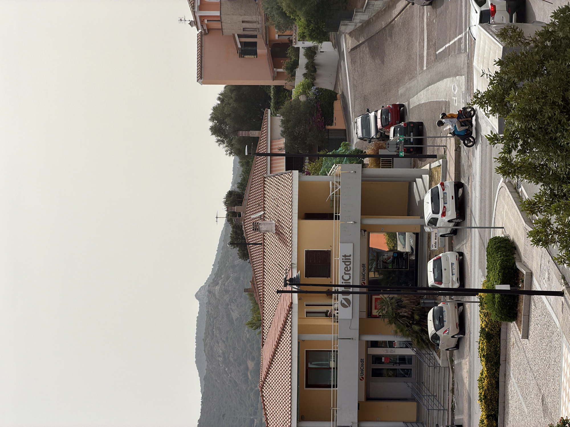 The image depicts a street scene in Olbia, Italy, on a hazy day. In the foreground, a two-story building with a yellow facade and red tiled roof houses a UniCredit bank on the ground floor, accessible by stairs. Several parked cars are visible, including a white Fiat 500, another white hatchback, and a dark SUV further up the street.

A paved road curves uphill, with more vehicles including a red hatchback and another white car. Two individuals are riding a scooter uphill, the rider wearing a helmet and the passenger a white shirt. The background features a residential building with an orange facade and a cluster of trees. In the far distance, a rugged, mountainous landscape is visible under an overcast sky. The scene appears to be a typical urban environment with commercial and residential elements.