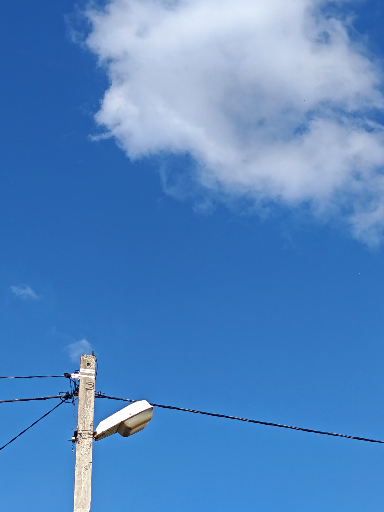 A concrete utility pole with a white streetlamp attached stands against a bright blue sky. Several black wires are strung between the pole and other unseen supports, disappearing into the upper left and right of the frame. A large, fluffy white cloud is visible in the upper right quadrant of the image. The streetlamp is elongated and appears to be made of white plastic or painted metal, with a translucent cover over the light source. The concrete pole is weathered and shows signs of age. The overall scene suggests an outdoor setting, likely in a residential or urban area, under clear weather conditions. The specific location in Mayrtup, Russia, is not discernible from the visual elements of the image.
