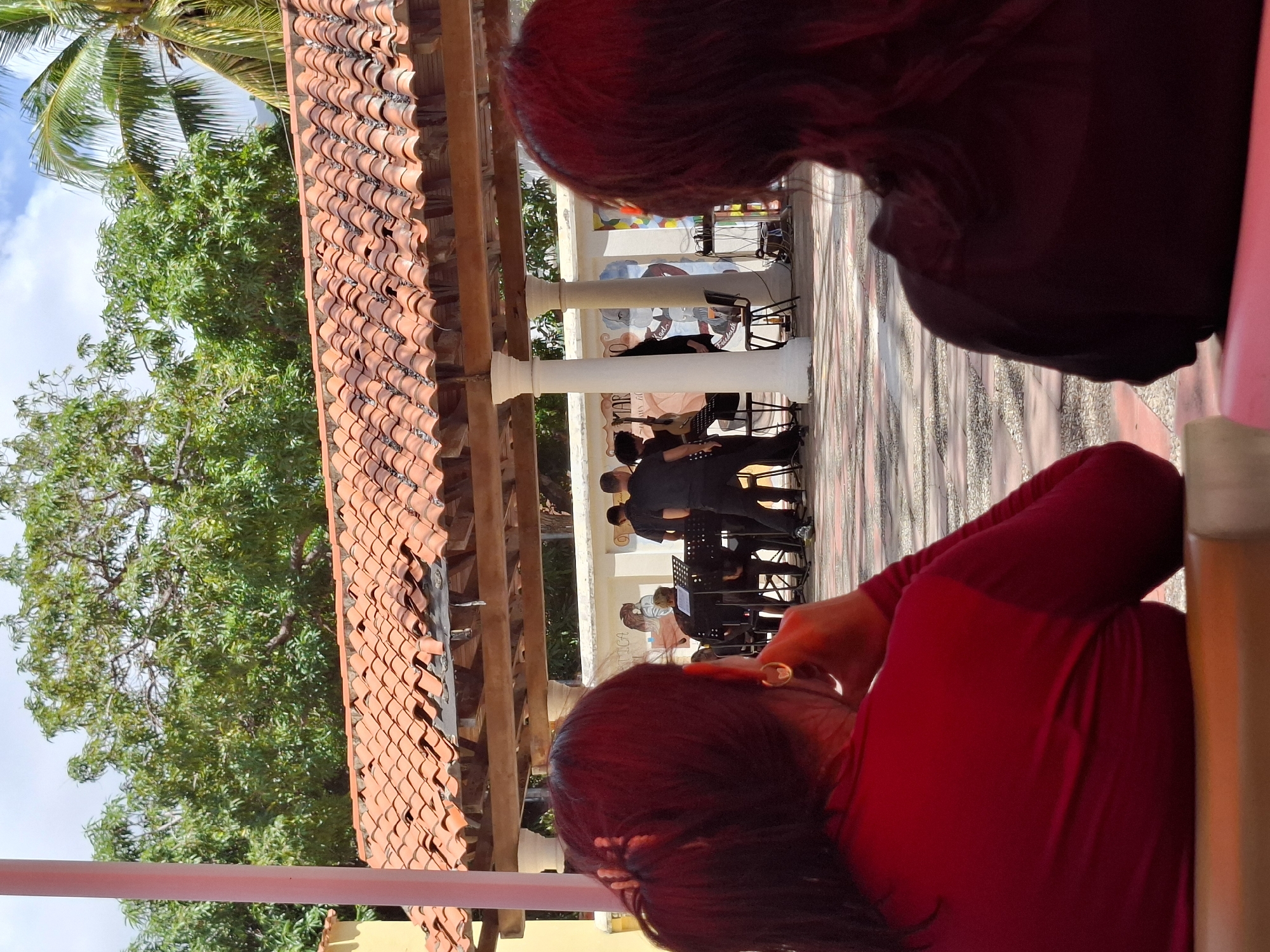 From a high-angle vantage point, the image captures an outdoor scene in El Valle del Espíritu Santo, Venezuela. Two individuals are prominently featured in the foreground. One is clad in red, with their head tilted back and their hand near their ear. The other person, positioned higher in the frame, is dressed in dark clothing.
In the mid-ground, a musical ensemble is visible. The musicians, dressed in black attire, are seated with their music stands.
The setting features terracotta tiled roofing, trees with green foliage, and a partially visible cloudy sky. The background includes a structure with white pillars and painted murals.