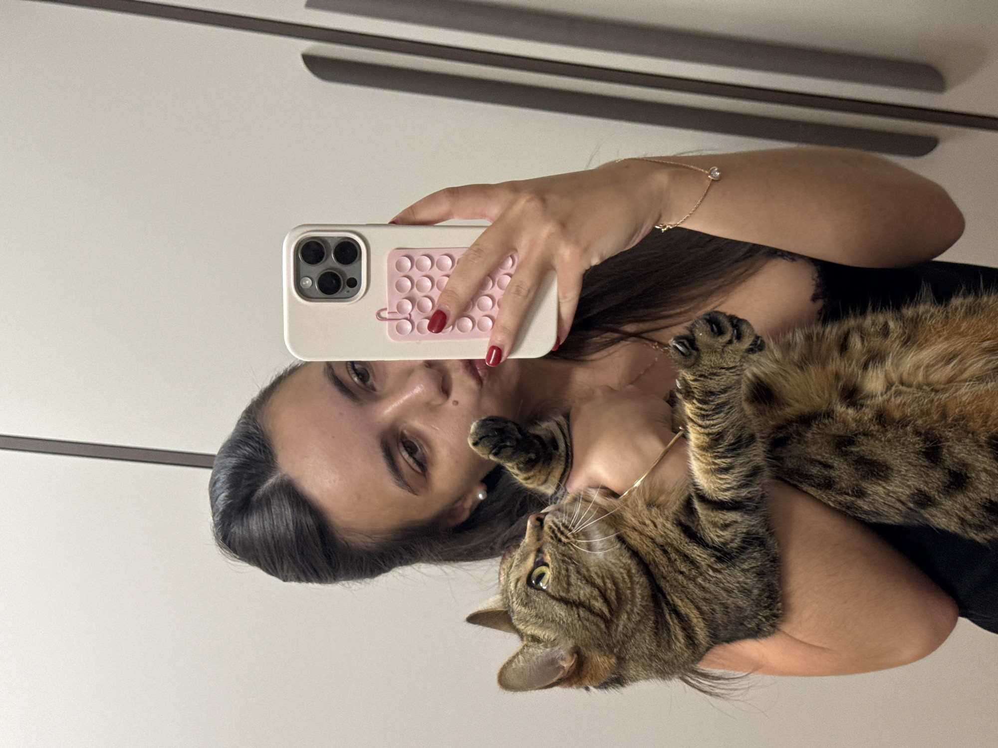 A young woman takes a mirror selfie, holding her cat. The woman has long dark hair and is wearing a black shirt and a delicate bracelet with a single diamond. She is also wearing red nail polish. Her cat is a tabby with brown and black stripes. The cat is lying on its back, being held by the woman. The background appears to be a wall with some horizontal lines, possibly indicating a piece of furniture or shelving. The lighting suggests it could be daytime or artificial indoor lighting. The woman's expression is neutral, while the cat looks up at the camera with wide, curious eyes. The image is taken from a slightly elevated angle, looking down at the subjects. There is no visible text in the image.