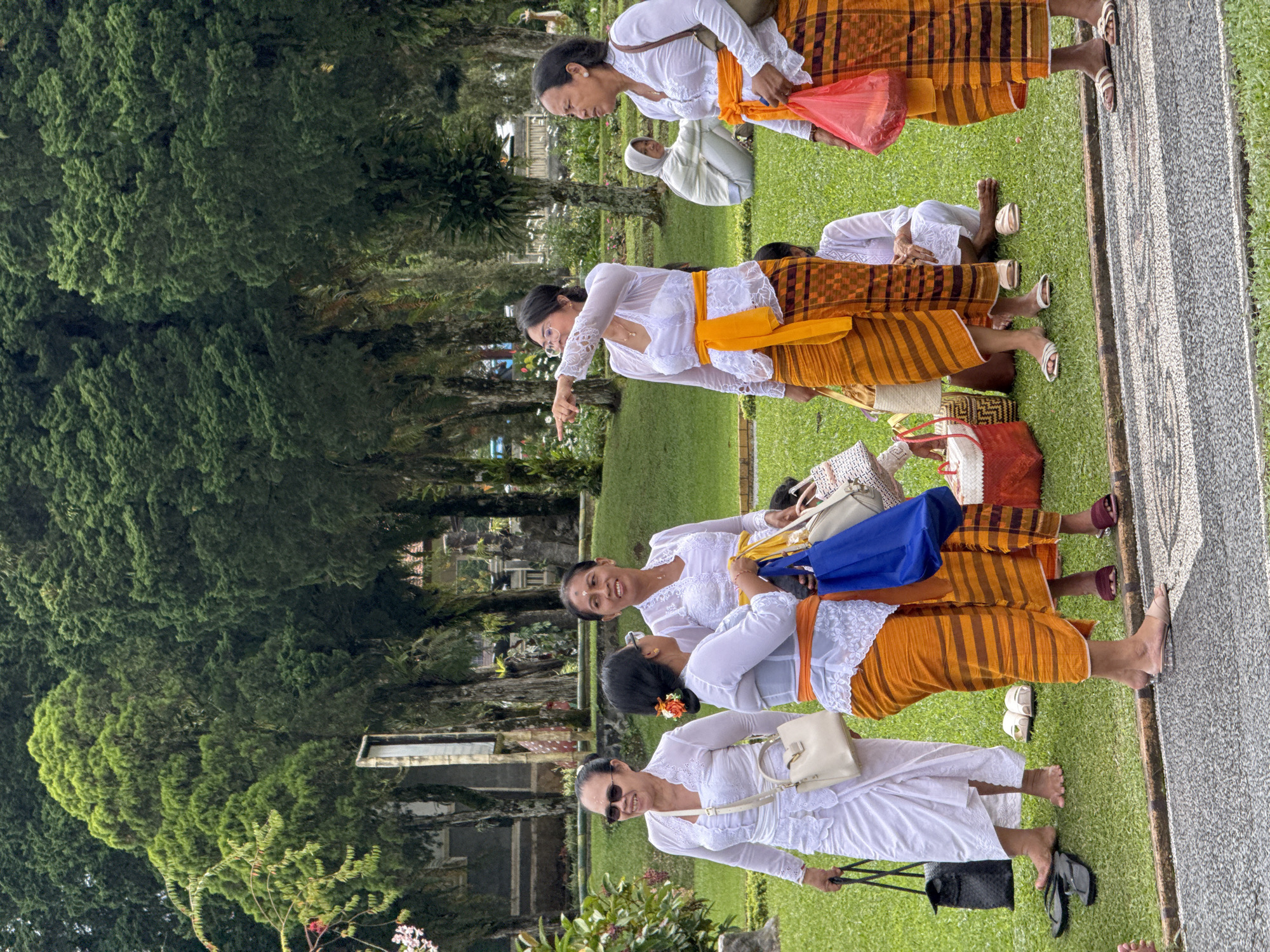 The image shows a group of women standing on grass next to a stone path in Baturiti, Indonesia. The women are wearing traditional Balinese attire, including white tops, colorful sarongs, and sashes. Some women are carrying bags. There are trees and greenery in the background.