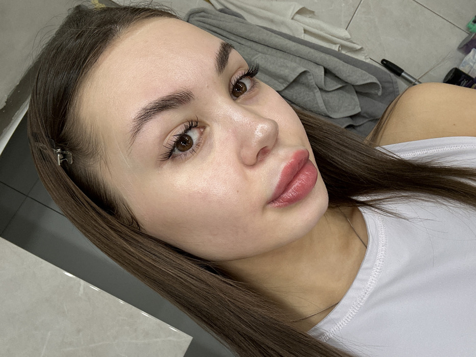 A young woman with light skin, brown hair, and brown eyes. She has full, pink lips and well-defined eyebrows. Her hair is parted to the side and held back with a small clip. She is wearing a white shirt, and the background appears to be a casual indoor setting with some items visible in the background. The lighting is bright, highlighting her features.
