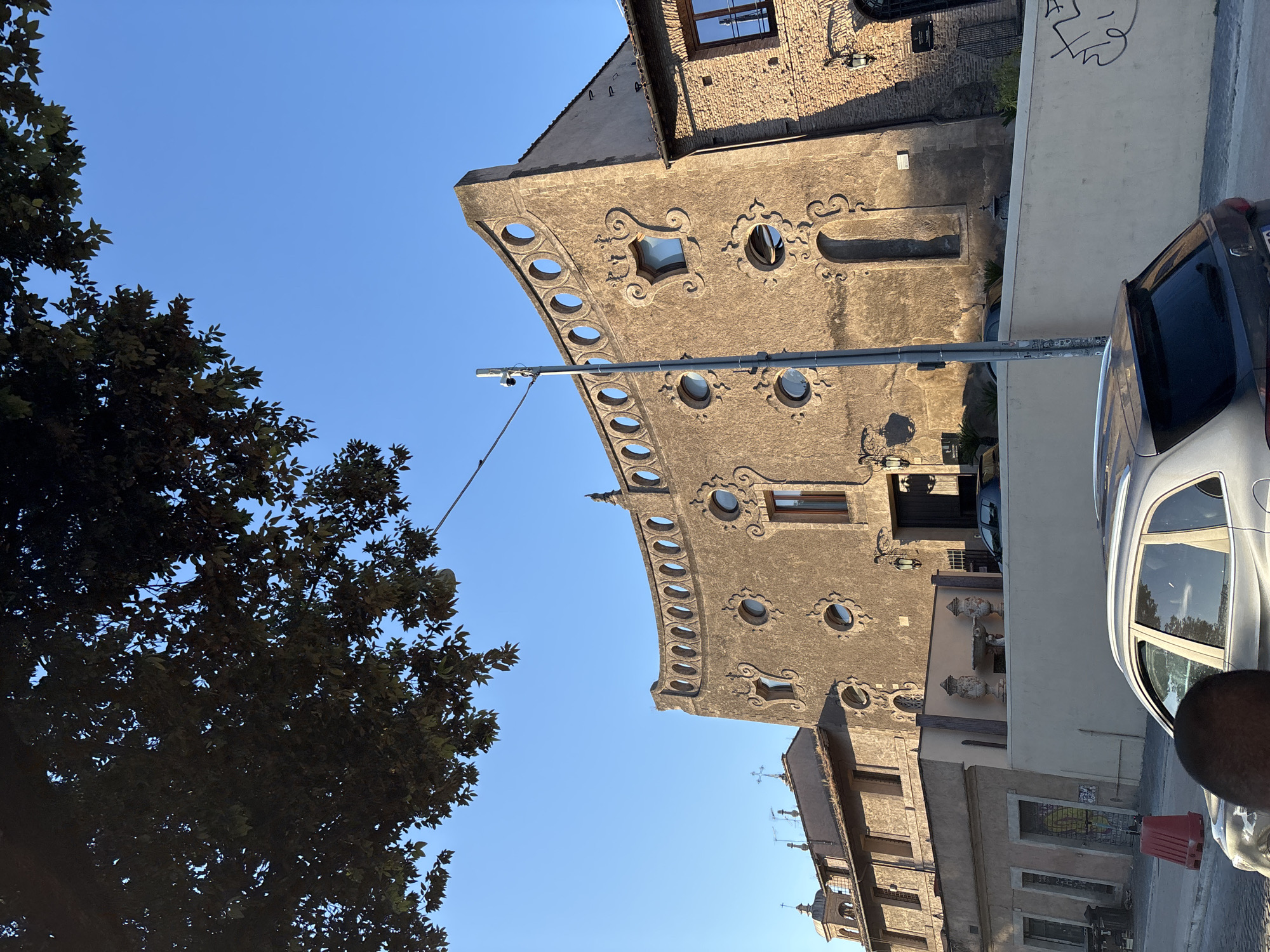 The image depicts a street in Rome, Italy, showcasing a unique building with distinctive architectural features. The building is characterized by a façade with circular windows and decorative scrollwork. A security camera is mounted on a pole extending towards the building. To the left, a tree with dense foliage partially obscures the blue sky. A silver car is parked on the right side of the street, with a portion of a person's head visible in the foreground. Other buildings line the street, one with a series of crosses and antennas on its roof. Graffiti is present on a wall adjacent to the car. The scene is bathed in sunlight, suggesting a clear day.