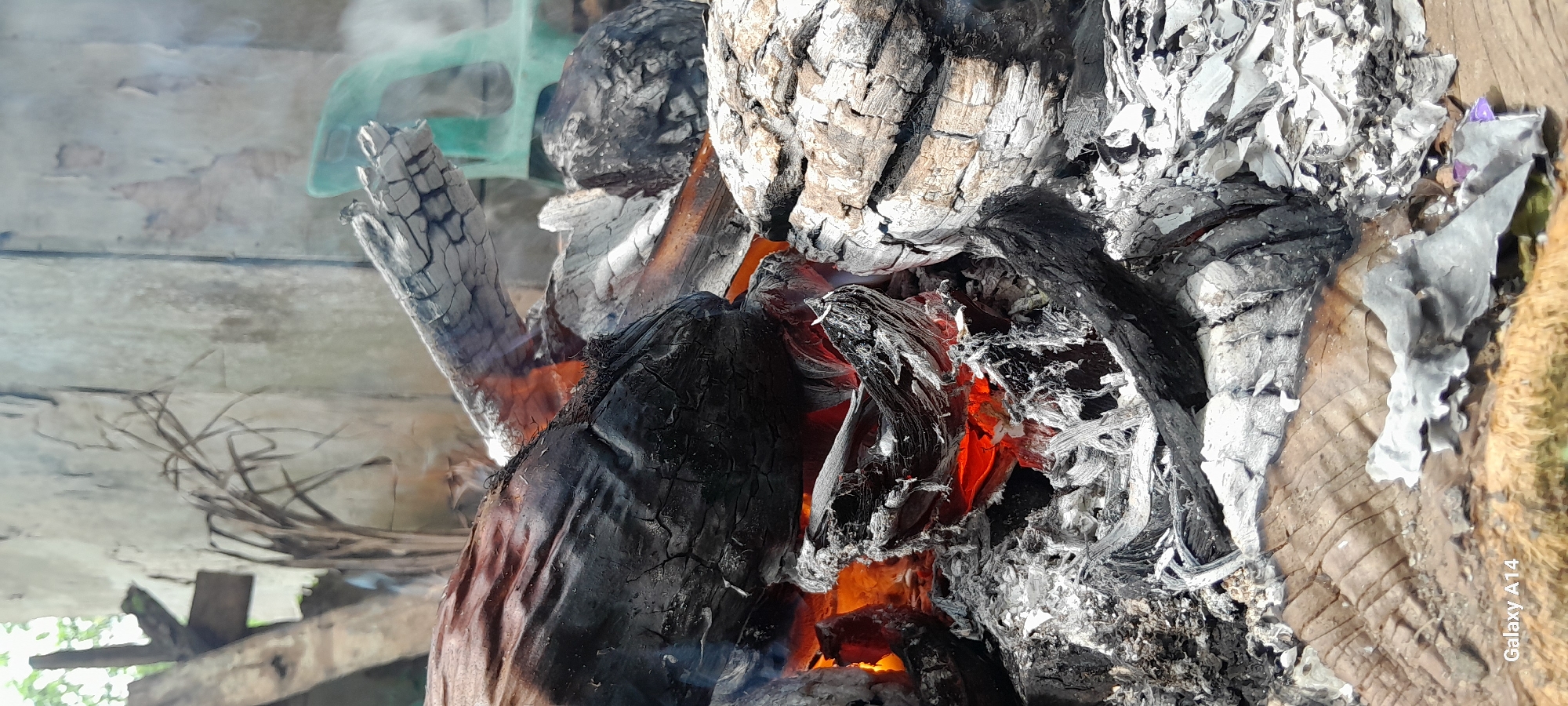 A close-up, eye-level, vertical shot shows a pile of burning wood embers and ash. The wood is charred black and gray with glowing orange embers visible in the center. Smoke drifts upwards from the fire. The background is blurred but appears to be an outdoor environment with concrete structures and greenery. The text "Galaxy A14" is visible in the bottom left corner. The location is stated to be Tanjong, Indonesia.