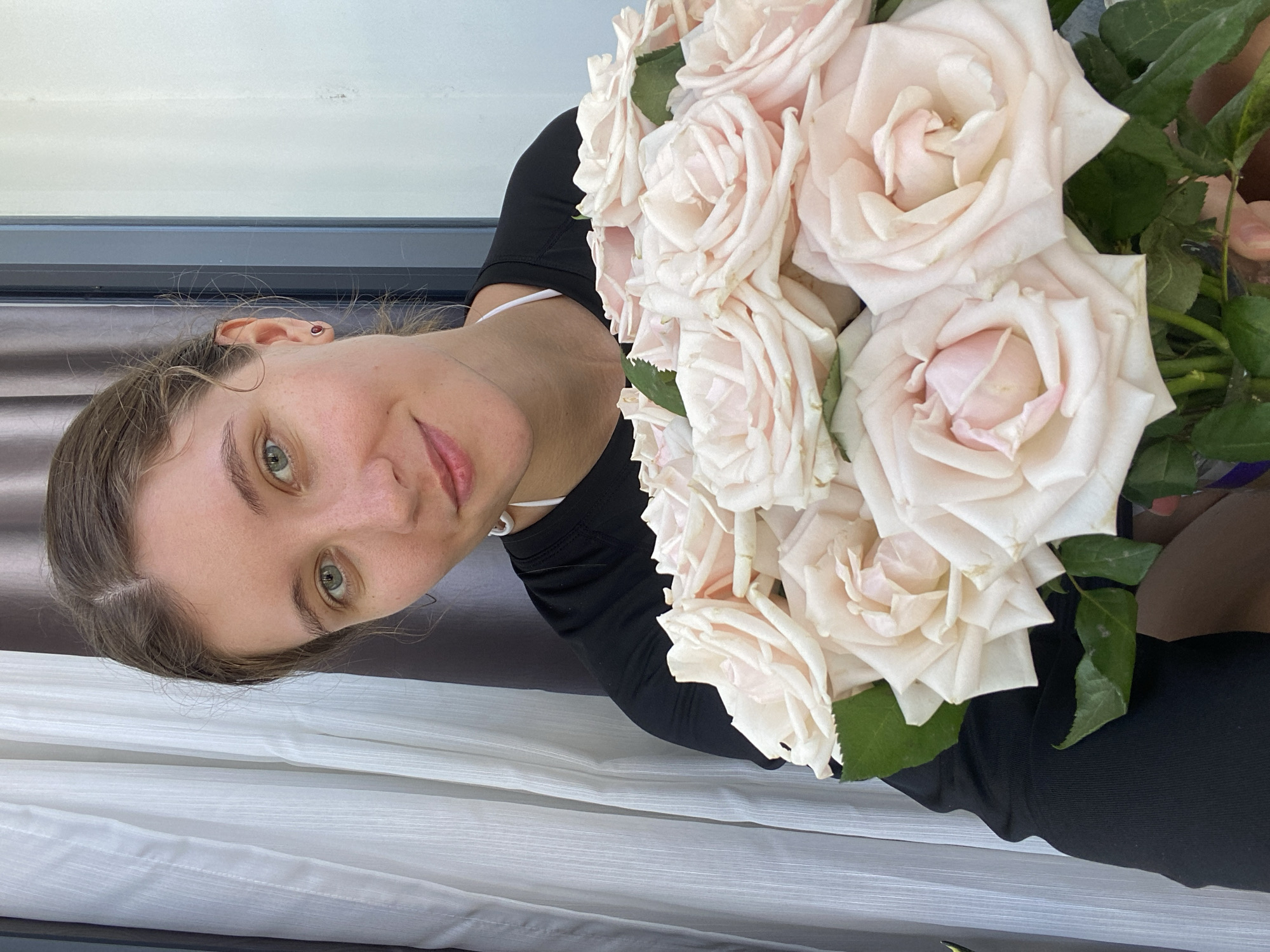 A young woman with light skin and green eyes, her hair tied back in a messy bun, is looking at the camera and smiling. She is wearing a black t-shirt and holding a large bouquet of light pink roses. The background appears to be indoors, with sheer white curtains on the left and a light-colored wall on the right. The scene suggests a personal moment, possibly a gift or a celebration, and the lighting indicates it's daytime. There are no visible elements that would place the scene specifically in Bac Nha Trang Ward, Vietnam; it is a generic indoor setting.