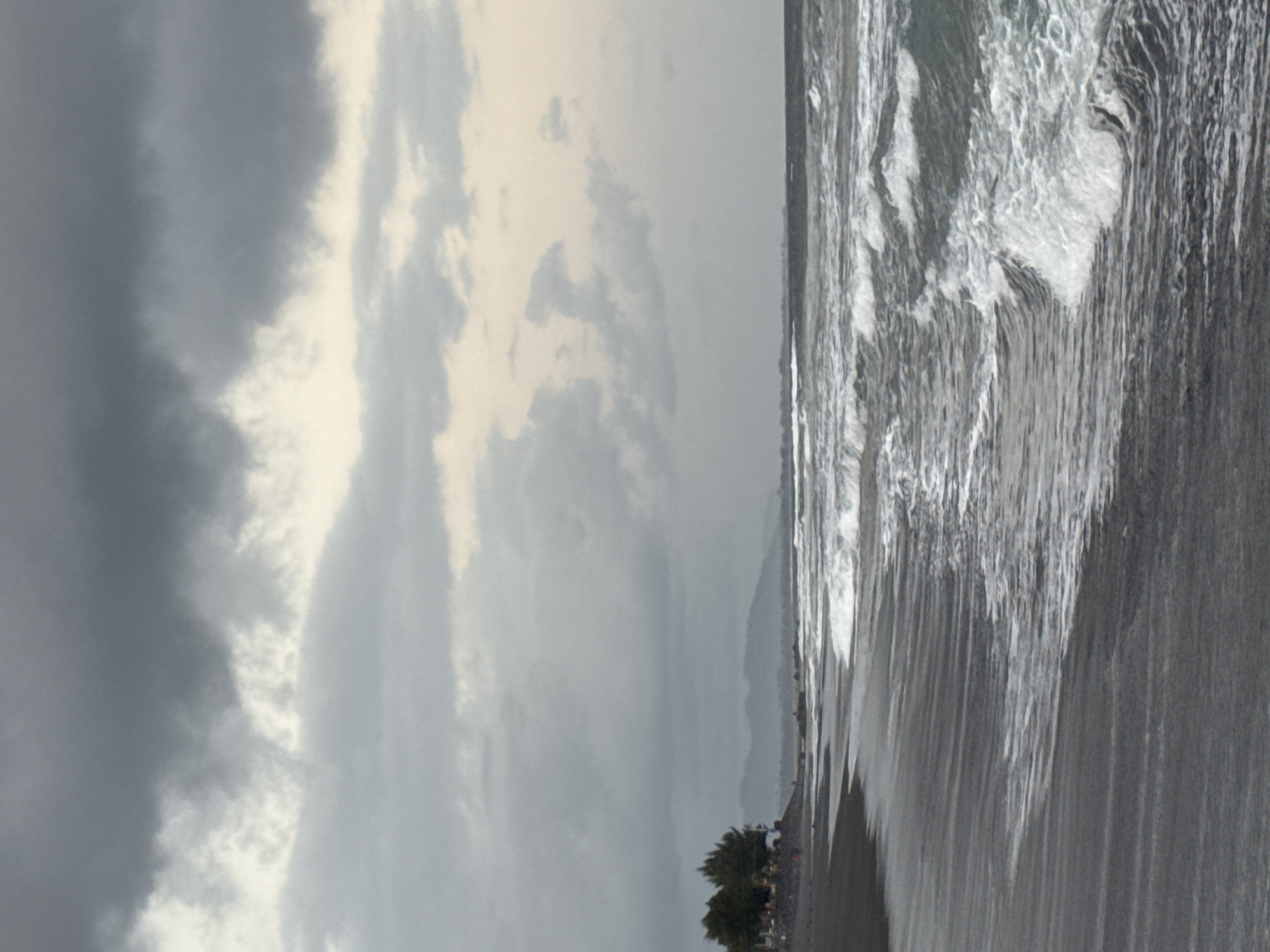 An overcast day at a beach in Blahbatuh, Indonesia, shows a dark sand shoreline meeting the ocean. Waves with white foam crest and break as they roll towards the shore. In the distance, a line of trees is visible along the coast, and further back, hazy mountains can be seen under the cloudy sky. A few indistinct figures are present on the beach.