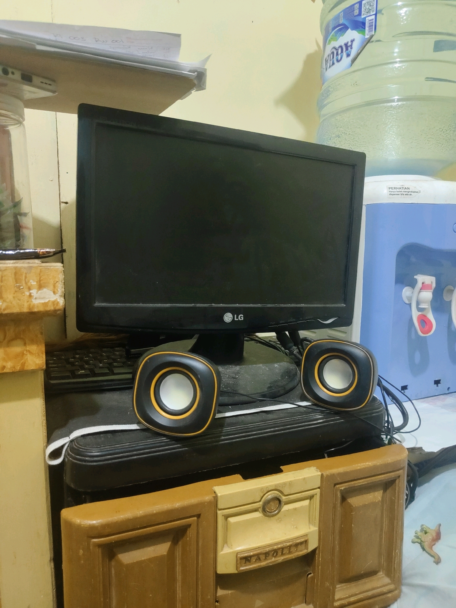In the city of Sukanagara, Indonesia, a scene unfolds within a room, likely a personal space, depicting a modest setup. Dominating the center is a black LG monitor, its screen dark, suggesting it is either off or displaying a black image. To its left and right, two small, black and orange speakers are positioned, their circular white cones visible. These speakers sit atop a black, multi-tiered object, possibly a computer tower or storage unit.
Behind the monitor and to the right, a large blue water cooler is visible, with a white dispenser spout featuring red accents. A clear plastic water bottle is perched atop the cooler, its label partially obscured but showing the "AQUA" brand. On the left of the scene, a glass jar with a white lid is present, its contents unclear. A shelf above this jar holds some papers, with writing visible on them.
Below the monitor and speakers, a black keyboard is partially visible. Further down, a tan-colored cabinet or drawer unit occupies the foreground, featuring decorative molding and a label that reads "NAPOLIT". A small, toy dinosaur, predominantly green and pink, lies to the right of this cabinet. The walls appear to be painted a light yellow. The overall impression is one of a functional, perhaps slightly cluttered, personal workspace or living area.