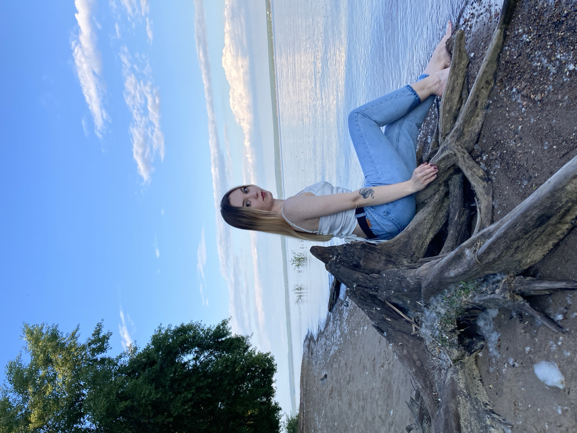 A young woman with blonde hair and red lipstick sits on the exposed roots of a large tree trunk on the shore of a large body of water. She is wearing a white top and blue jeans. The water is calm and reflects the blue sky with scattered white clouds. A large green tree stands to the left of the frame, casting a shadow on the sandy shore. The area appears to be a natural, undeveloped setting.