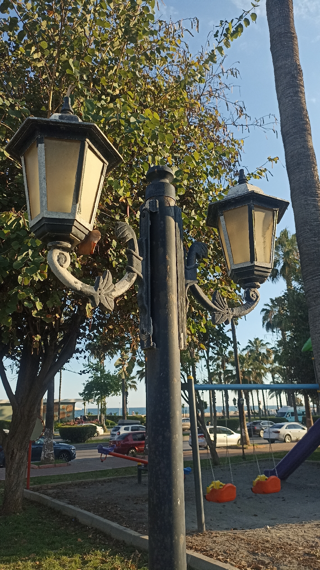 The image captures a sunny day in a park area located in Yenişehir, Turkey, with the sea visible in the background. The primary subject is a large, black street lamp with two ornate lanterns, positioned prominently in the foreground and slightly off-center to the left. The lanterns have a classic design with yellowed glass panes and decorative metalwork, including scroll and leaf motifs on their supporting arms.
Behind the street lamp, a dense, leafy green tree with round leaves and elongated seed pods fills much of the upper left and central frame. To the far right, the textured trunk of a tall palm tree is visible. In the midground, a children's playground is present, featuring two orange swings and part of a purple slide structure on the right side, along with a red seesaw. The ground in the playground appears to be a mix of dirt and sand.
Beyond the playground and a low concrete border, a road runs horizontally across the scene. Several cars of various colors (including blue, red, and white) are visible on the road, some parked, some possibly moving. A lush strip of greenery, including more palm trees, lines the far side of the road. In the distant background, the tranquil blue sea meets a clear, light blue sky. The soft, directional sunlight suggests either morning or late afternoon. No people are visible in the scene, and there is no visible text.