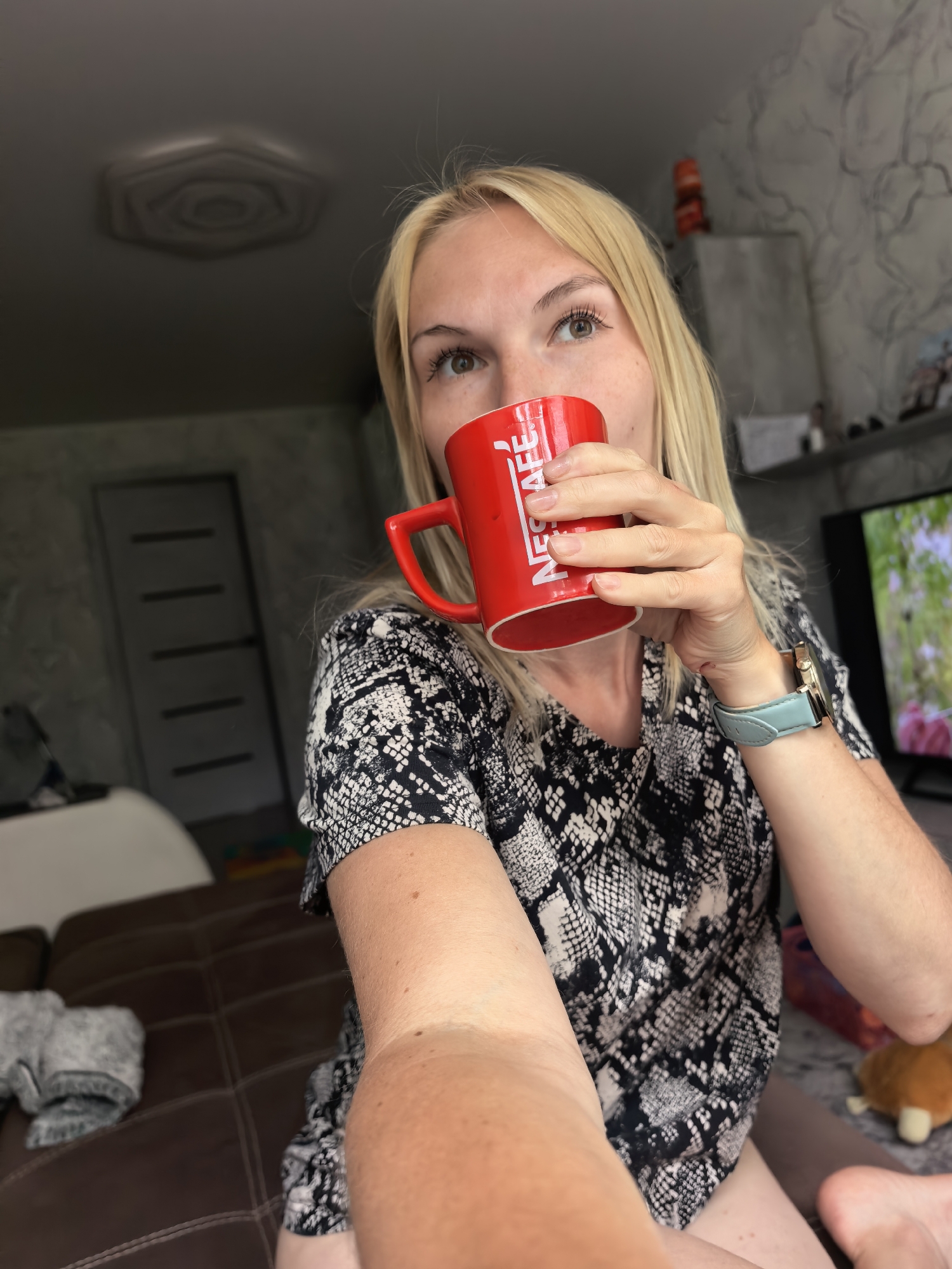 A blonde woman drinks from a red Nescafé mug. She is wearing a black and white patterned shirt and a light blue watch. The scene appears to be indoors, possibly in a living room. A brown sofa is visible to the left, and a television screen is on the right. The ceiling features a textured design. The overall lighting suggests it is daytime. The specific location within Sterlitamak, Russia, cannot be determined from the image alone.