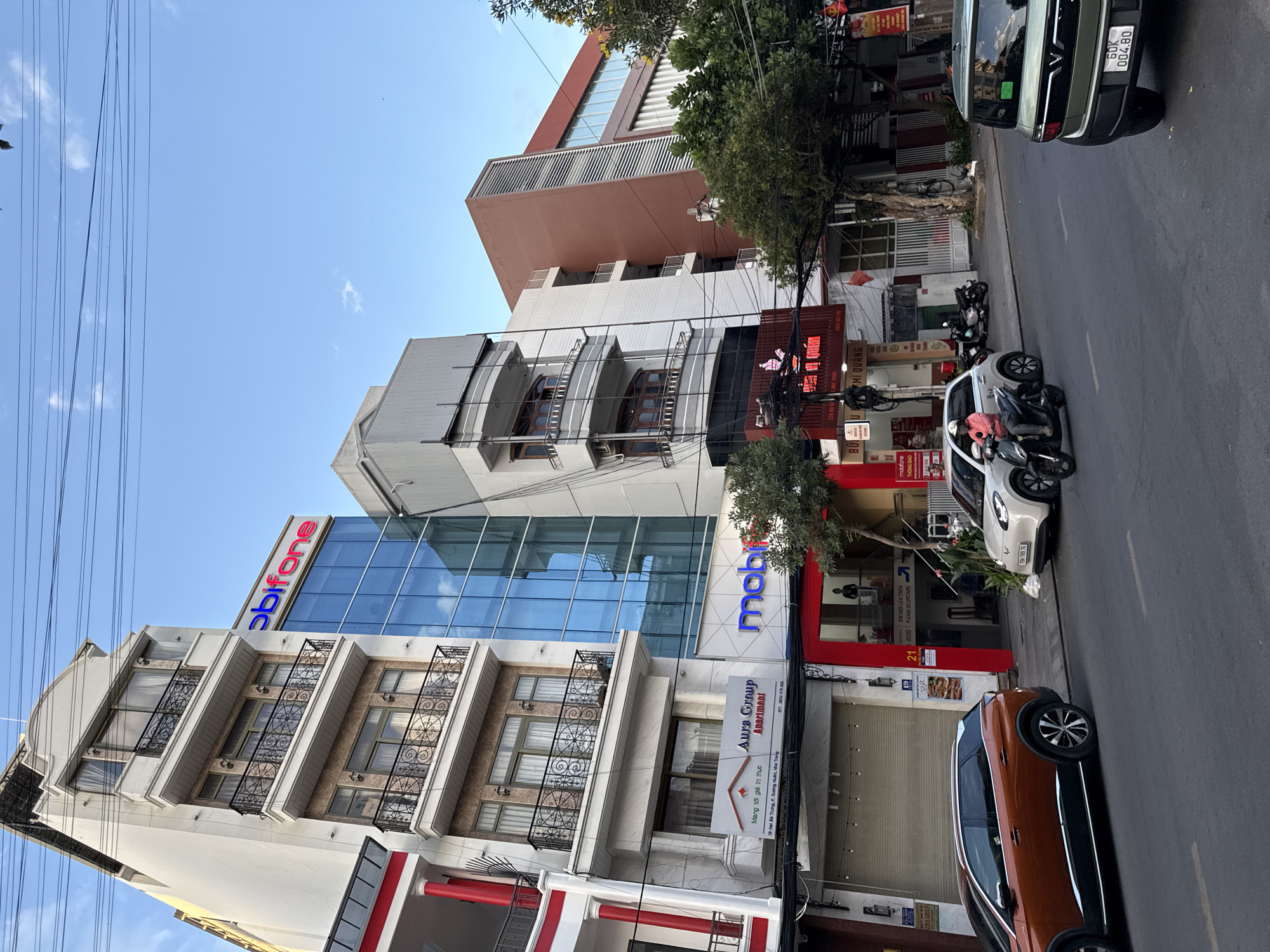 This image captures a street scene in Nha Trang, Vietnam, focusing on the architecture and a few vehicles. On the left, a multi-story building with white and beige facades features balconies with ornate black railings. Adjacent to it, a modern building with a blue glass facade displays the "mobifone" logo prominently in red and blue neon. Further to the right, another building with a reddish-brown facade shows a more contemporary design with vertical elements.
In the foreground, a street runs horizontally across the image. Two cars are parked or driving on the street: an orange SUV on the left and a white electric car on the right. A person on a scooter is visible, navigating the street. Signs with Vietnamese text are present on the buildings, indicating businesses. The sky is blue with a few scattered clouds, and overhead, power lines are strung across the scene. The overall impression is that of a typical urban street in a Vietnamese city, with a mix of older and newer construction, commercial activity, and vehicular traffic.
