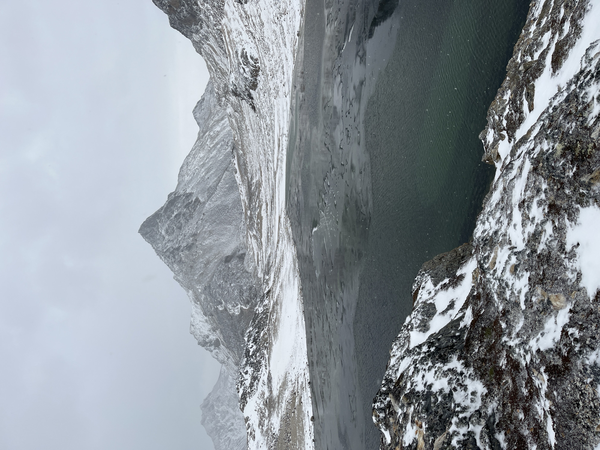 The image shows a mountainous, snowy landscape. In the foreground is a rocky cliff covered with snow and lichen. Adjacent to the cliff is a body of water, possibly a lake or fjord, with a dark green hue. In the background, there is a towering mountain range, also covered in snow, and shrouded in mist or fog. The sky is overcast and gray. There is no indication of any presence of people, subjects, objects, or interactions. It is impossible to confirm the exact location within Ushuaia, Argentina, based on the provided image alone.