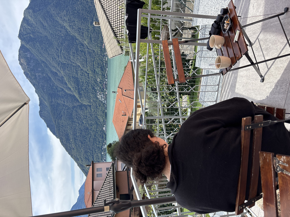 A single individual is seated with their back to the camera on an outdoor terrace in Nesso, Italy, overlooking Lake Como and forested mountains under a partly cloudy sky. The person has dark, curly hair tied back in a bun and wears a black long-sleeved top, sitting on a wooden slatted chair with metal supports. A set of keys rests on an adjacent wooden chair to the left in the foreground. To the right of the seated individual, a small wooden folding table with metal legs holds two light-colored ceramic mugs (one containing a spoon), a black ashtray filled with cigarette butts, a white cigarette, a black cylindrical object, and a folded dark fabric item. The terrace floor is gray tile, and a metal railing with geometric patterns separates the immediate area from the view. Beyond the railing, residential buildings with orange and gray tiled roofs are visible, descending towards the turquoise water of Lake Como. Across the lake, a distant settlement is discernible at the base of expansive, green, tree-covered mountains that dominate the background. A large white patio umbrella is partially visible in the upper left of the frame.