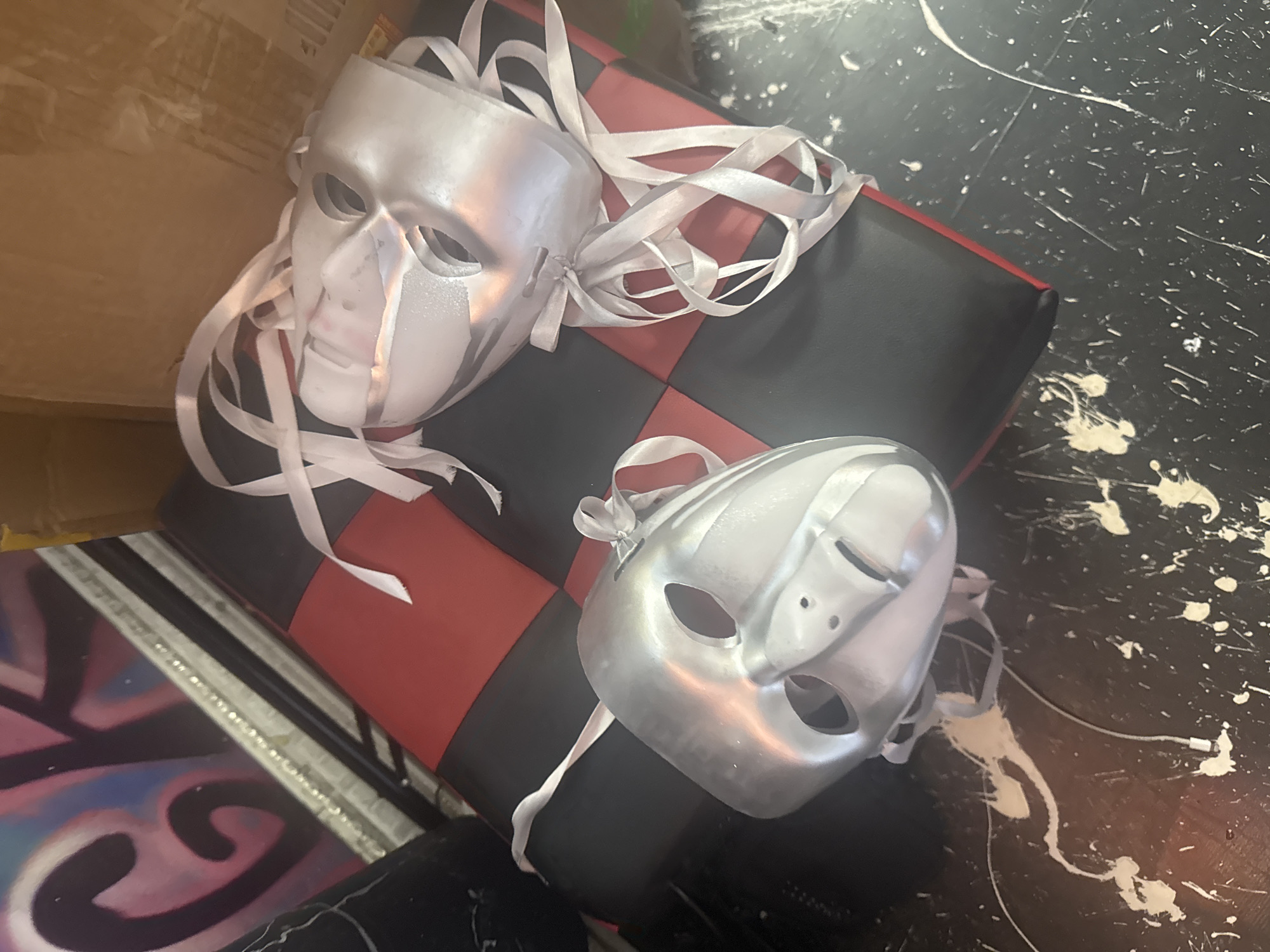 In Komsomolsk-on-Amur, Russia, the image depicts two silver masks with white ribbon ties resting on a black and red checkered surface, possibly a cushion or mat. The masks are oriented with one facing left and the other facing right. The background includes what appears to be a cardboard box and a framed piece of art with graffiti featuring pink, purple, and blue lettering. The surface beneath the masks shows splatters of white paint or a similar substance. The lighting is uneven, casting some reflections on the masks.