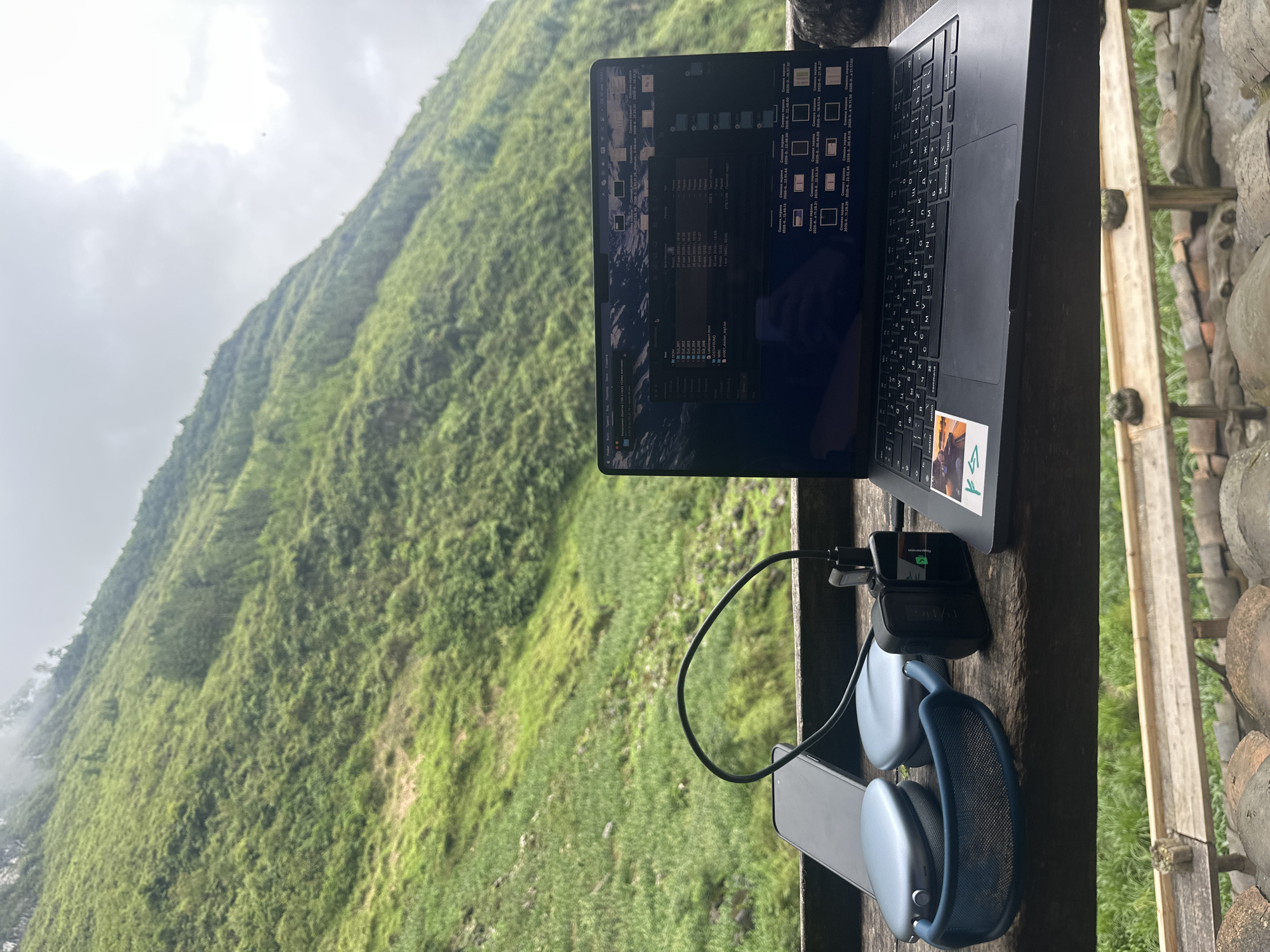 A laptop computer, possibly a MacBook, is open on a wooden surface with a view of a verdant, steep hillside in the background. The vegetation on the hillside appears dense and lush, possibly mossy, and partially obscured by mist or low clouds. To the right of the laptop is a pair of blue AirPods Max headphones resting on top of a grey smartphone and a black charging adapter with a short cable connected. The laptop screen displays a file explorer window with multiple files and folders. The wooden surface appears to be part of a structure with additional wooden elements to the right and stone materials in the background. The scene is located in the Yên Minh Commune, Vietnam.