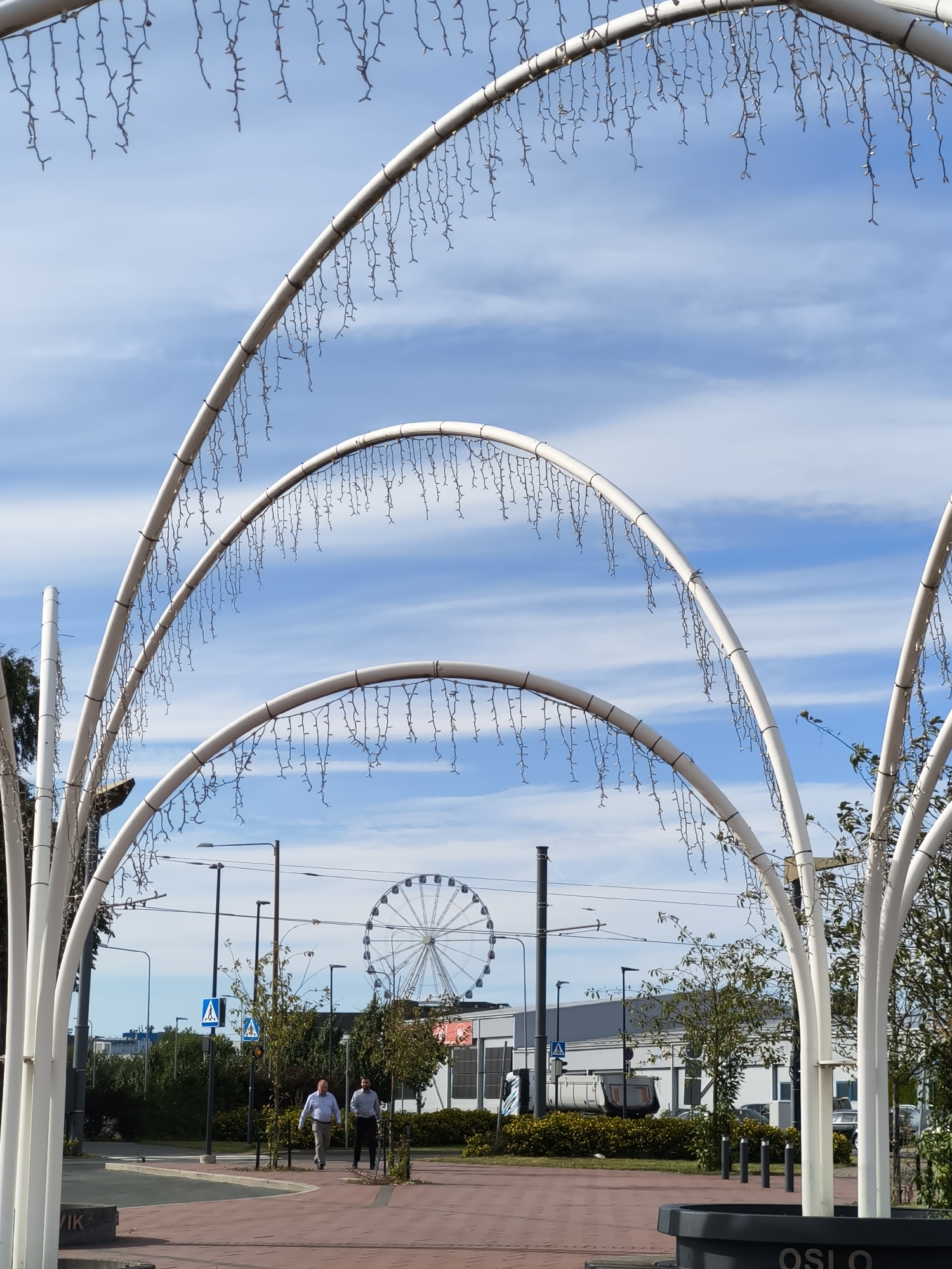 The image depicts an outdoor scene in Tallinn, Estonia, featuring a large Ferris wheel in the background. In the foreground, a white, arched structure adorned with numerous small, hanging lights creates a tunnel-like effect. Two men in business attire are walking on a red brick pathway. The pathway is bordered by green bushes and trees. Streetlights and pedestrian crossing signs are visible. The sky is blue with scattered white clouds. The overall impression is of a public space, possibly a park or a promenade, decorated for an event or a festive occasion.