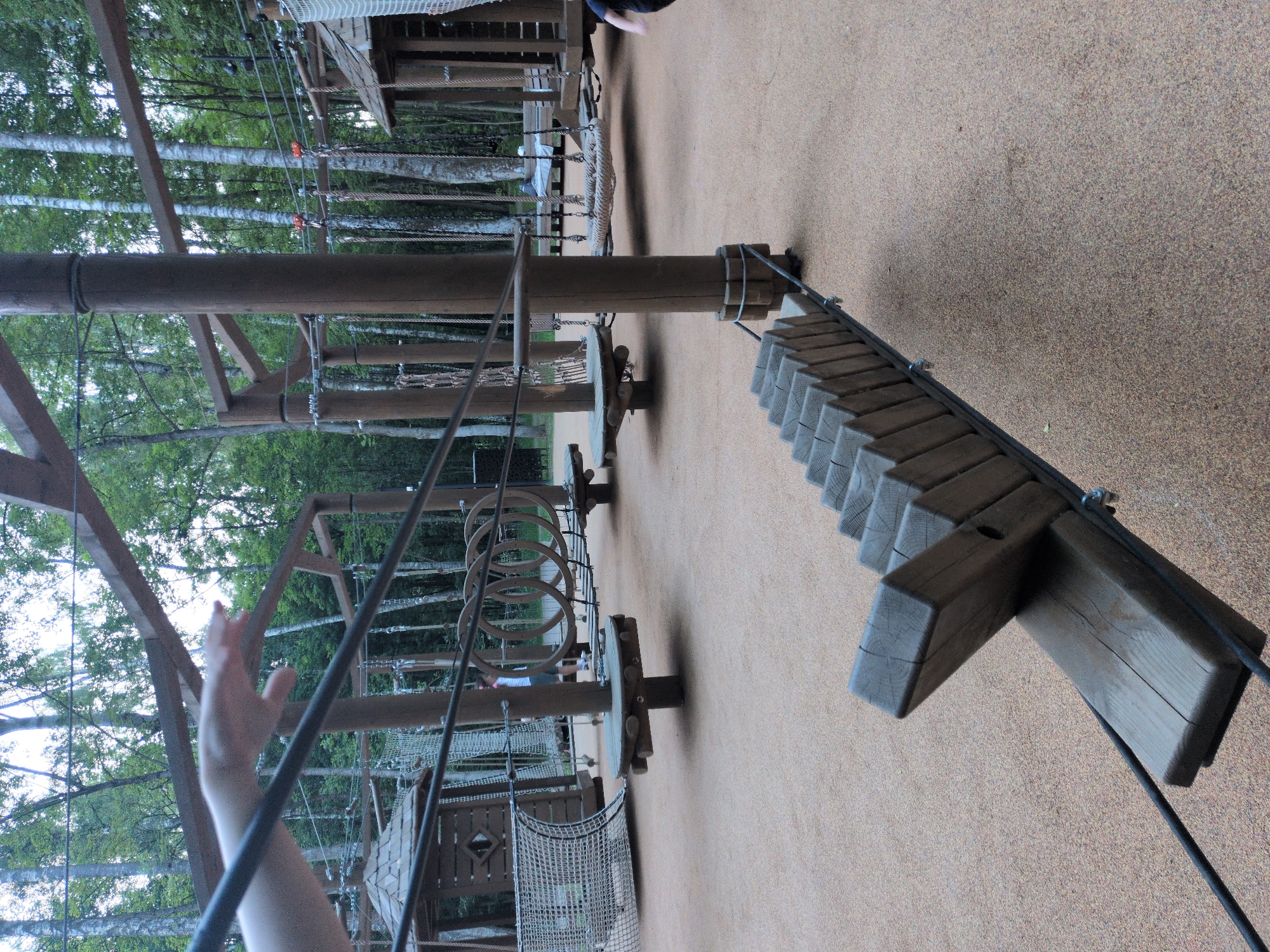 The image depicts an outdoor adventure park, likely located in Pelikhovo, Russia, featuring a challenging rope course and wooden obstacles. In the foreground, a person's hand is visible, reaching out and blurred due to motion, suggesting they are participating in or about to participate in the course. Dominating the mid-ground is a unique wooden bridge-like structure made of angled planks suspended by cables, designed to test balance. Behind this, the course continues with various elements, including wooden platforms, rope bridges, and net enclosures, all supported by sturdy wooden posts. In the background, a dense forest of tall trees creates a natural, shaded environment. A few other individuals are partially visible, one standing near a wooden structure and another in the far right, in motion. The ground is covered with a soft, light-colored material, typical of adventure park safety surfacing. The overall scene conveys an atmosphere of outdoor recreation and physical challenge.