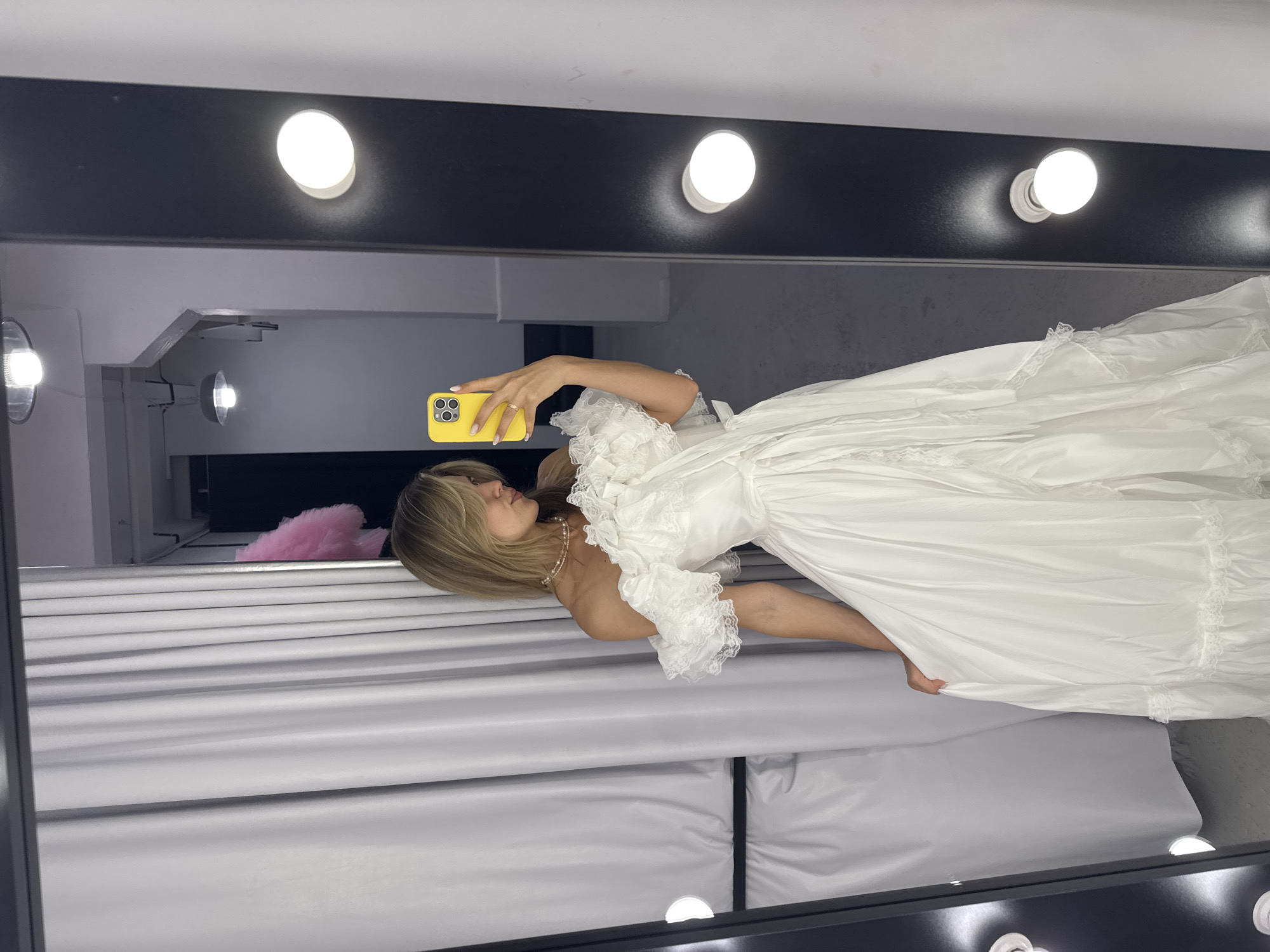 A woman with blonde hair, wearing a white off-the-shoulder gown with lace trim and a pearl necklace, is taking a selfie in a mirror. She is holding a yellow smartphone. The background includes white curtains, a black vanity mirror with light bulbs, and overhead lights. The setting appears to be a dressing room or a retail space.