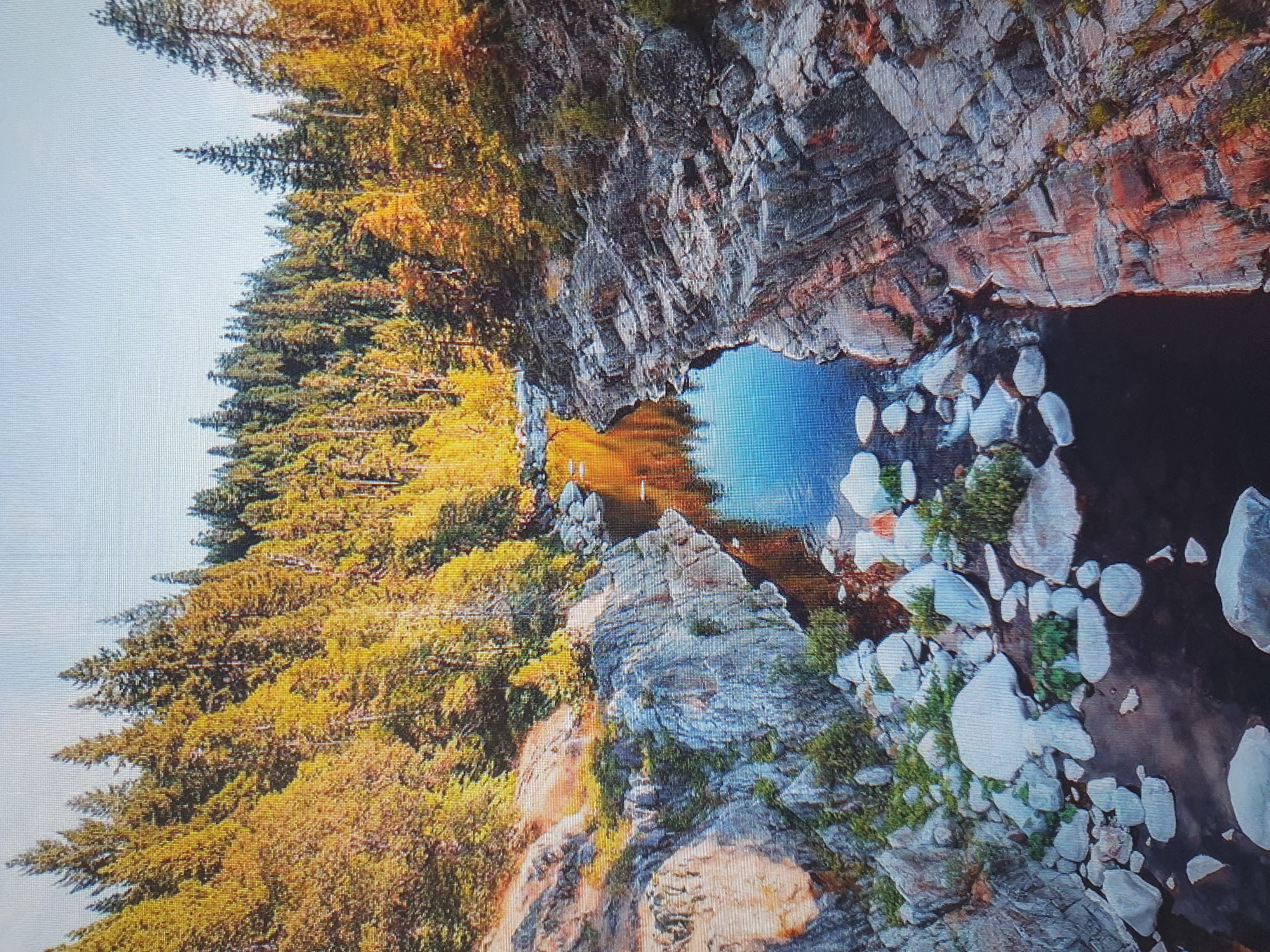The image displays a natural landscape featuring a river flowing between rocky banks, surrounded by a dense forest. The trees on the slopes exhibit vibrant autumn colors, with a mix of green and golden foliage. The river is calm, reflecting the sky and the surrounding trees. Large, smooth rocks are scattered along the riverbed and on the banks. The overall scene evokes a sense of wilderness and the beauty of nature during the fall season.
This image does not depict any location in the city of Tashkent, Uzbekistan. The landscape shown is characteristic of a mountainous or forested region with a river, not an urban environment.